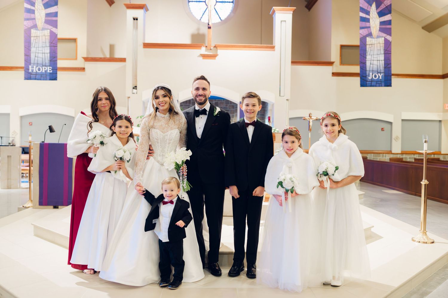 Wedding party portrait with bride, groom, bridesmaids, and flower girls inside decorated church.