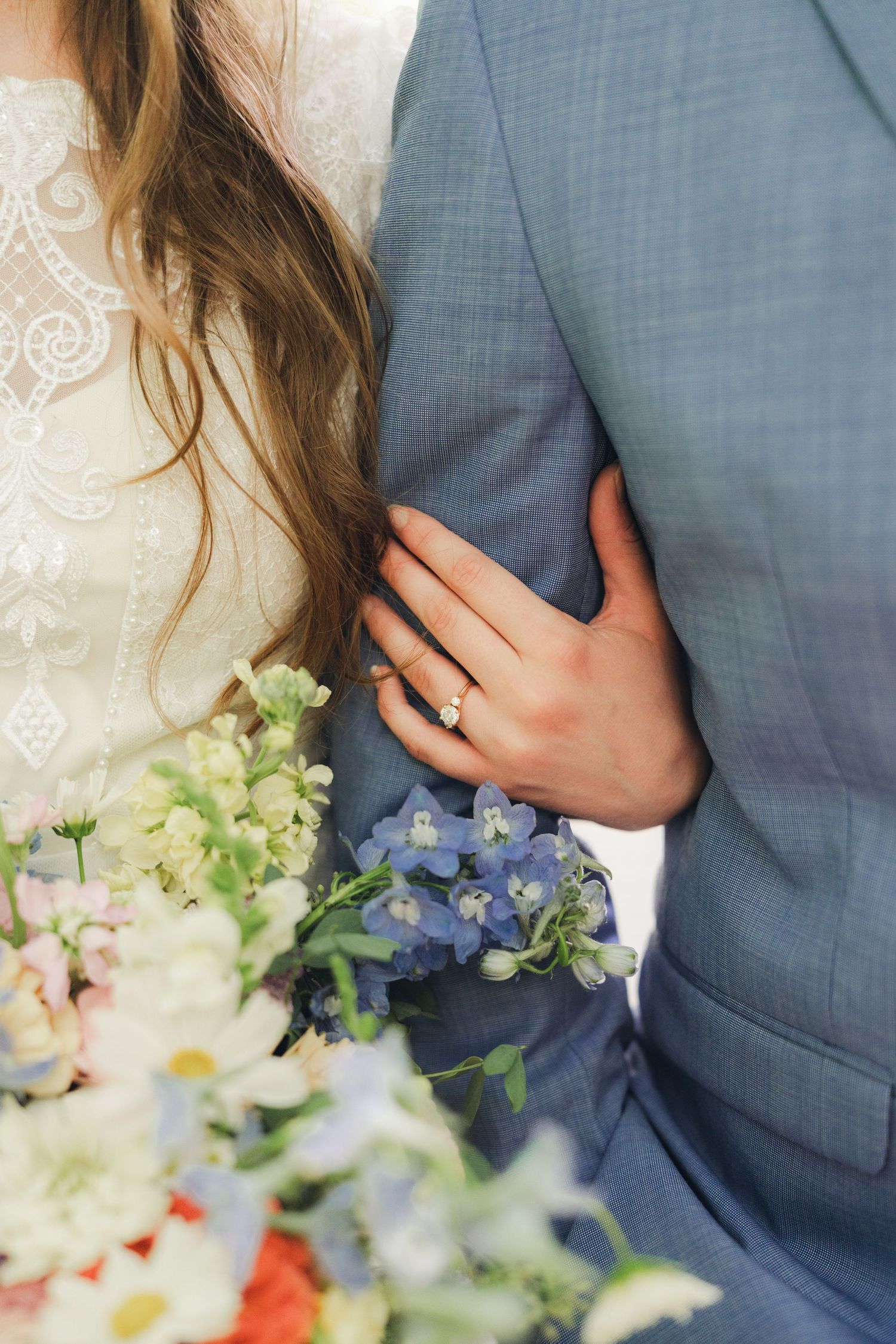 Bride in lace dress holding colorful bouquet with diamond ring visible while embracing groom in blue suit.
