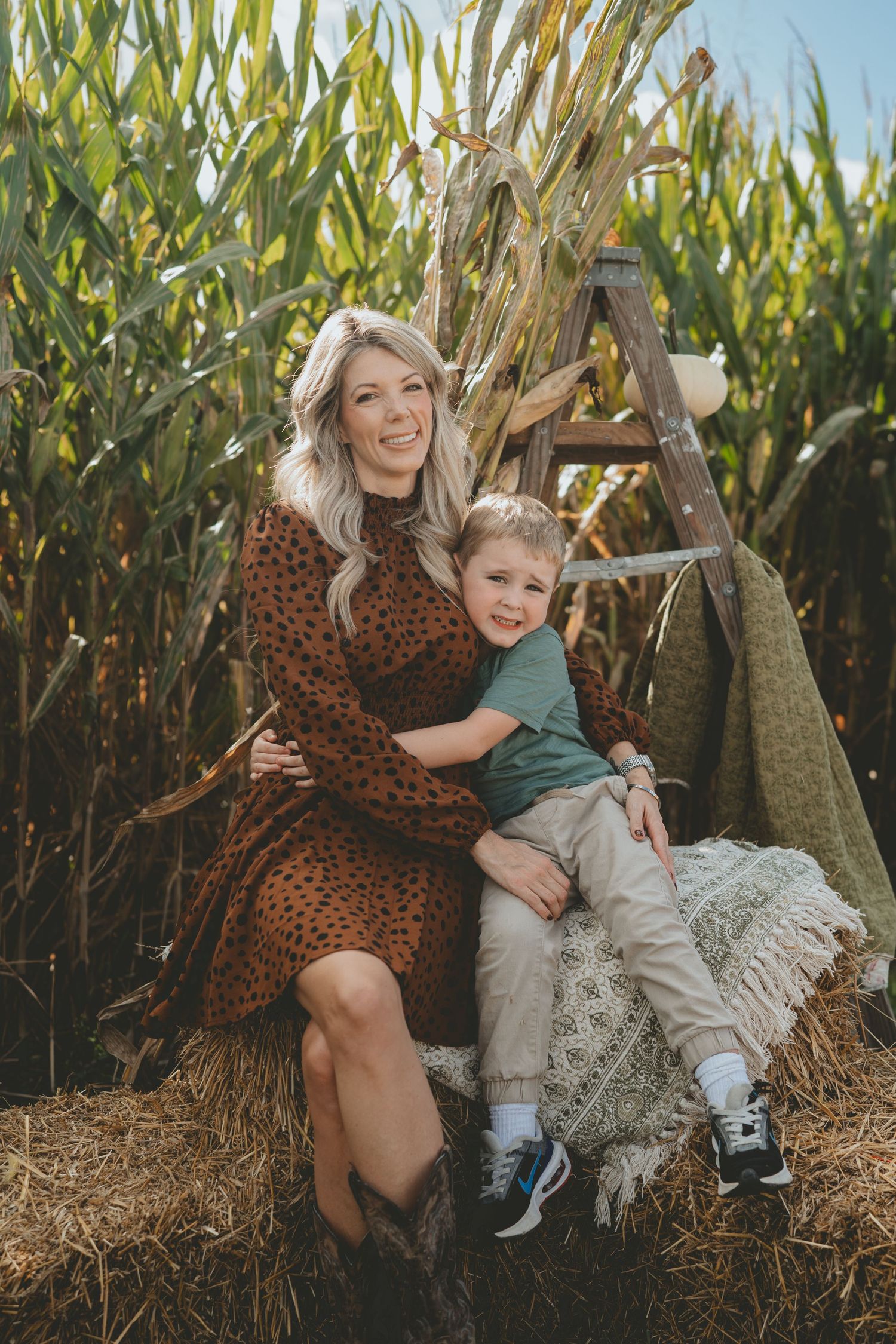 Blonde woman in leopard dress hugging young son on hay bale by cornfield.