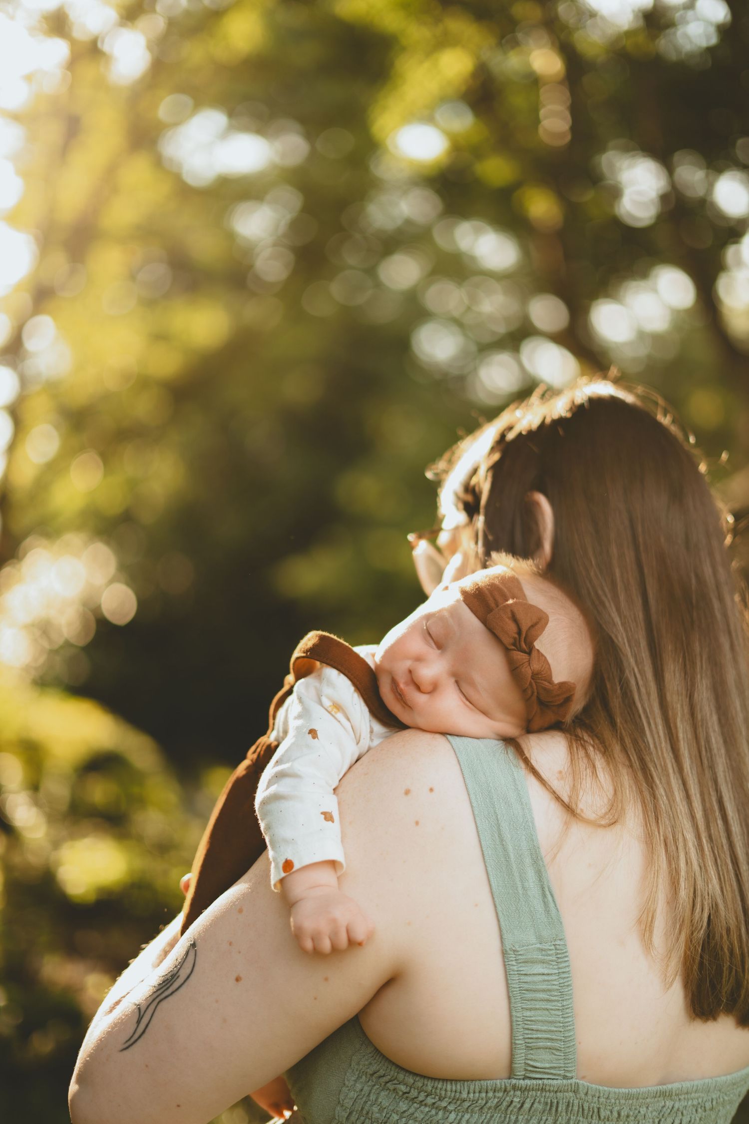A mother holds her sleeping newborn baby over her shoulder in a sun-drenched outdoor garden setting.