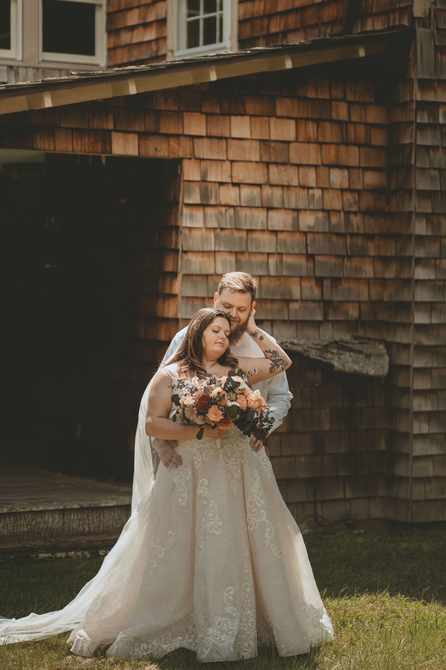Bride and groom embrace tenderly in front of a rustic cedar shingle building on their wedding day.