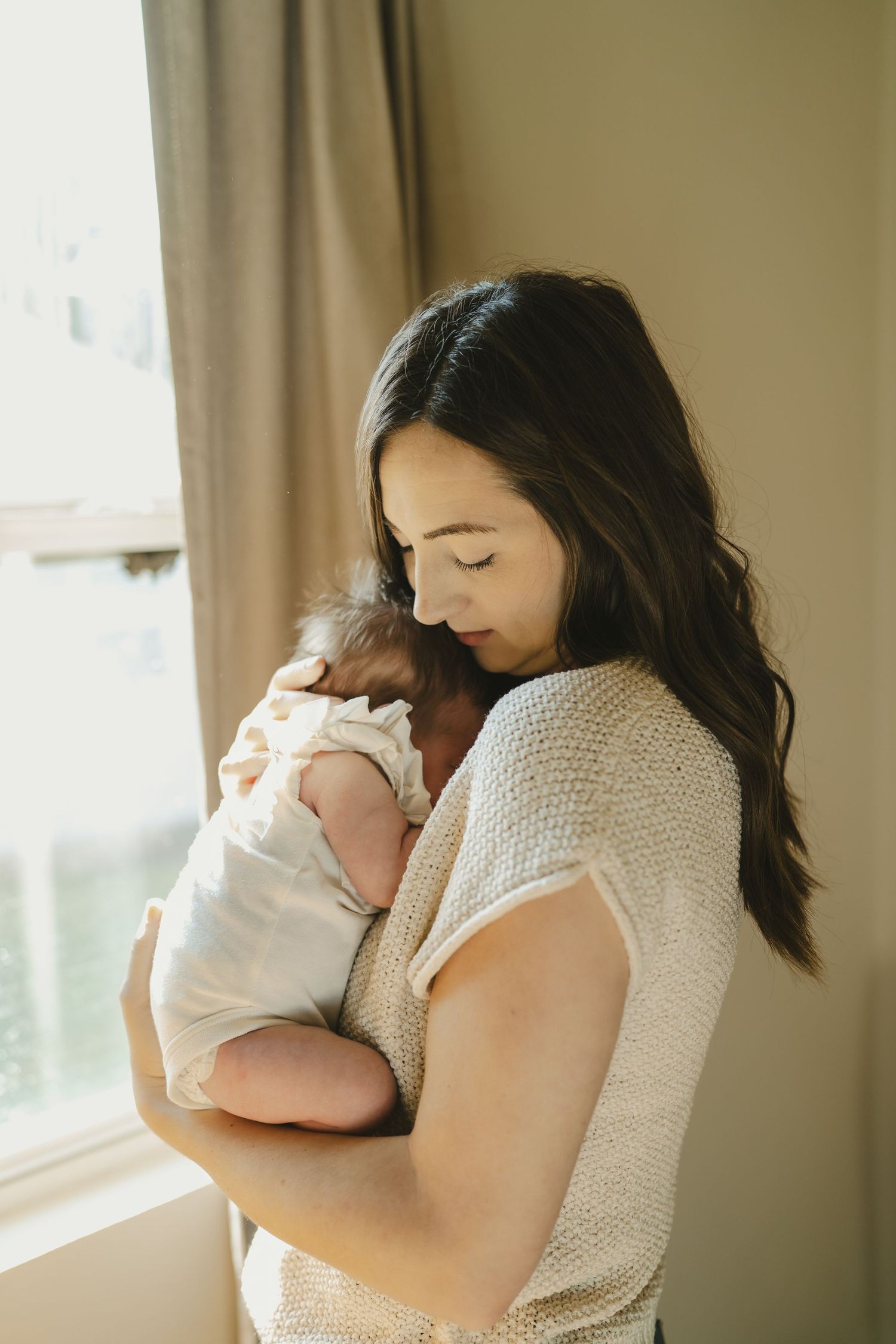Dark-haired mother standing by window holding swaddled newborn baby close to her chest.