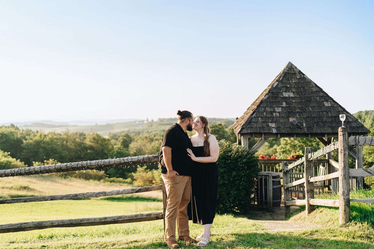 Couple sharing a romantic kiss near a wooden gazebo with scenic countryside views on a sunny day.