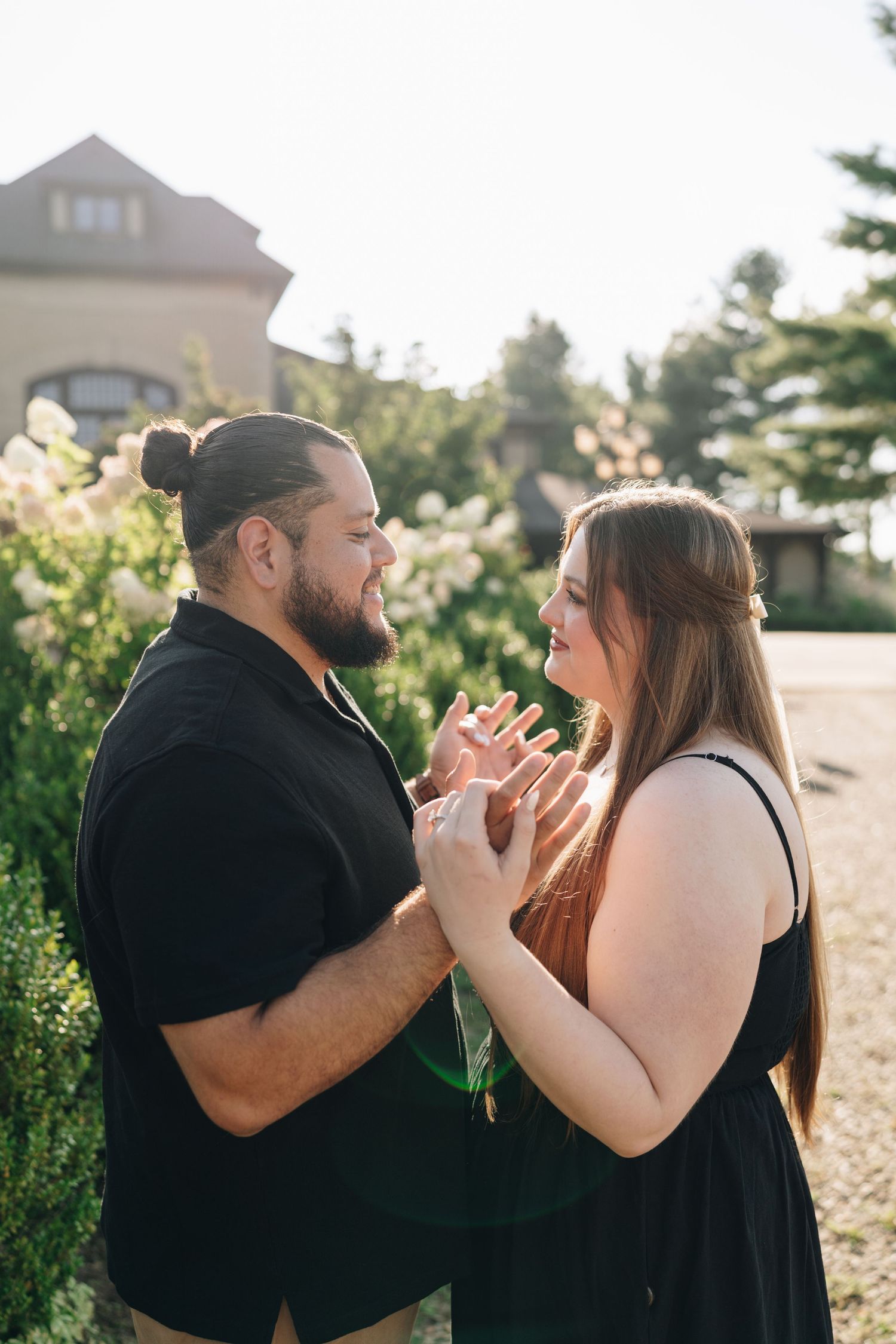 Couple holding hands and facing each other lovingly in front of white flowering bushes at golden hour.