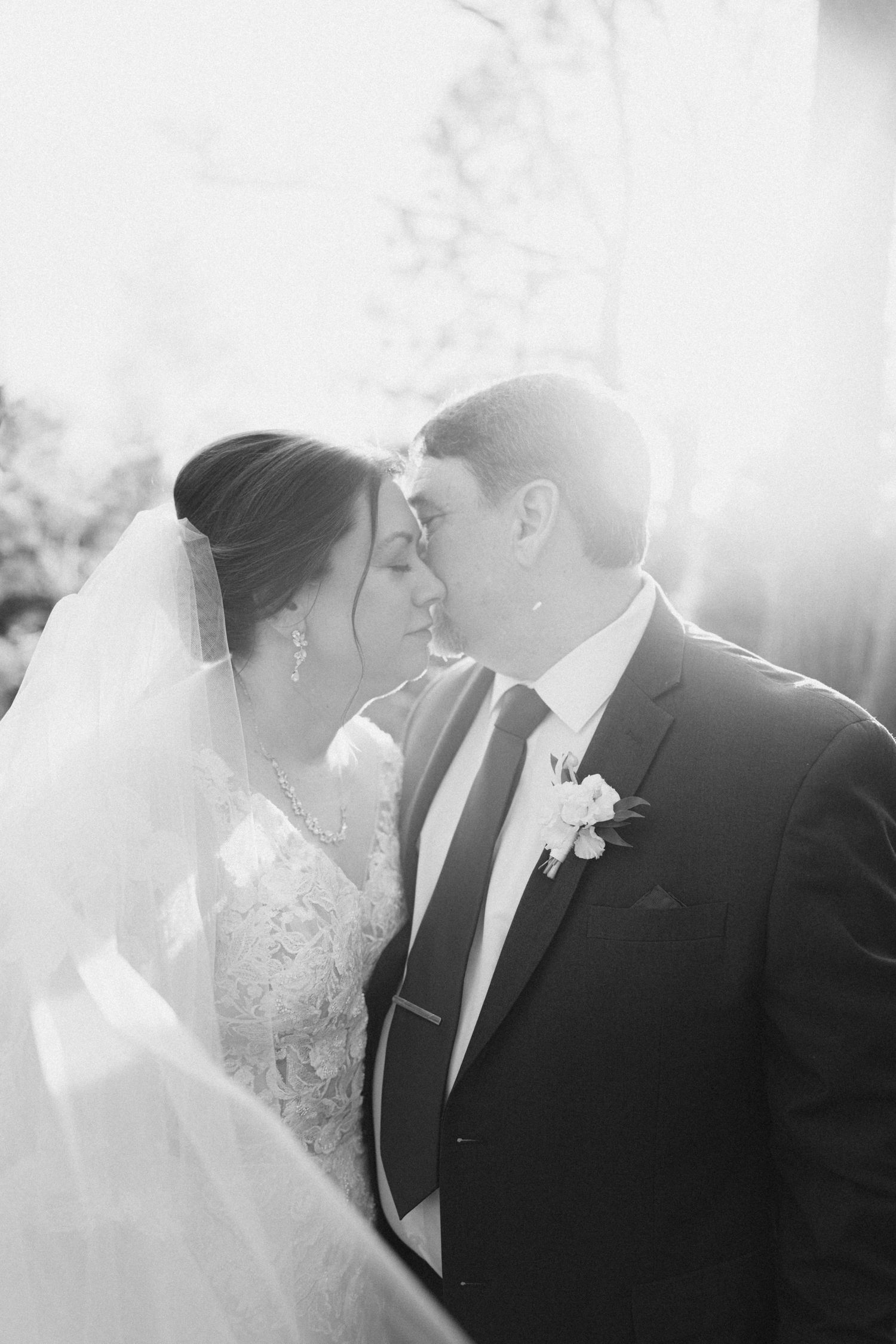 Black and white romantic portrait of bride and groom touching foreheads under flowing veil in sunlight.