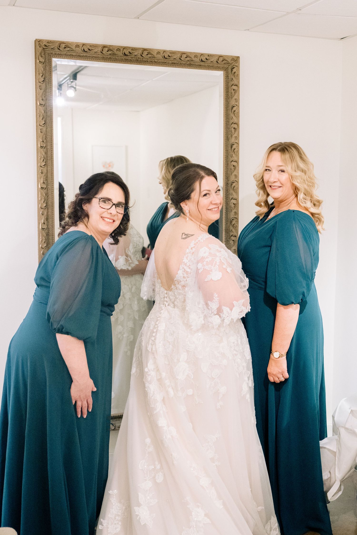 Bride in lace wedding gown poses with two bridesmaids in teal dresses in front of an ornate gold mirror.