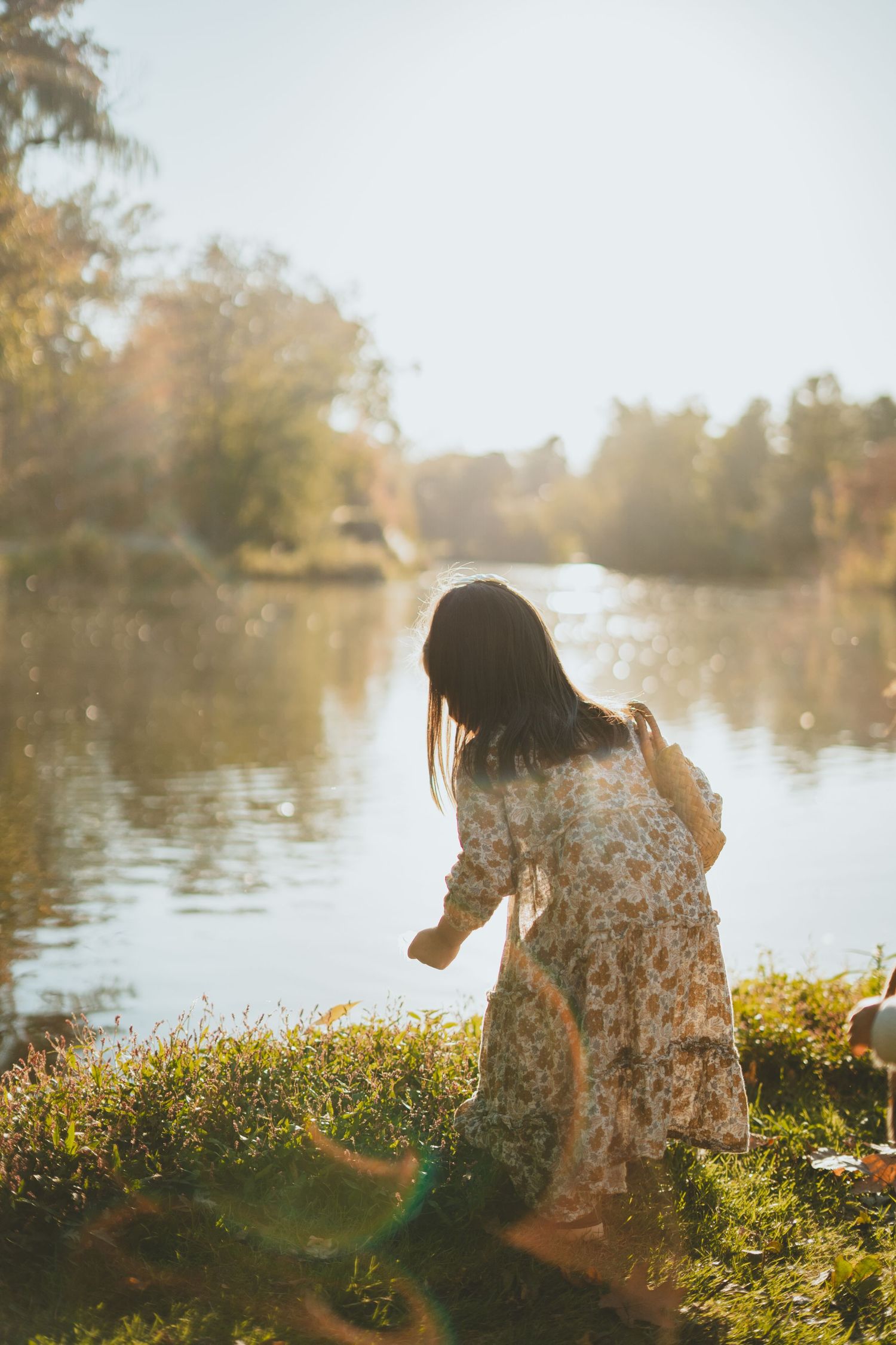 Woman in floral dress standing by a sunlit lake in autumn park.