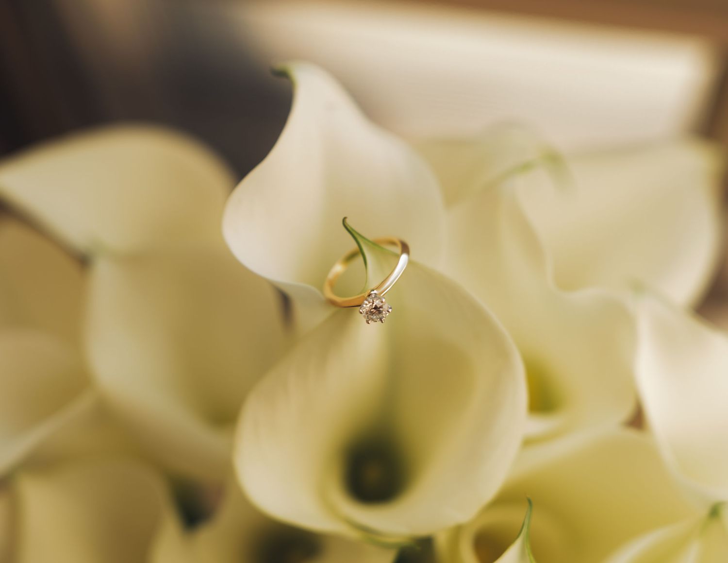 Diamond solitaire engagement ring nestled among white calla lily wedding bouquet flowers.