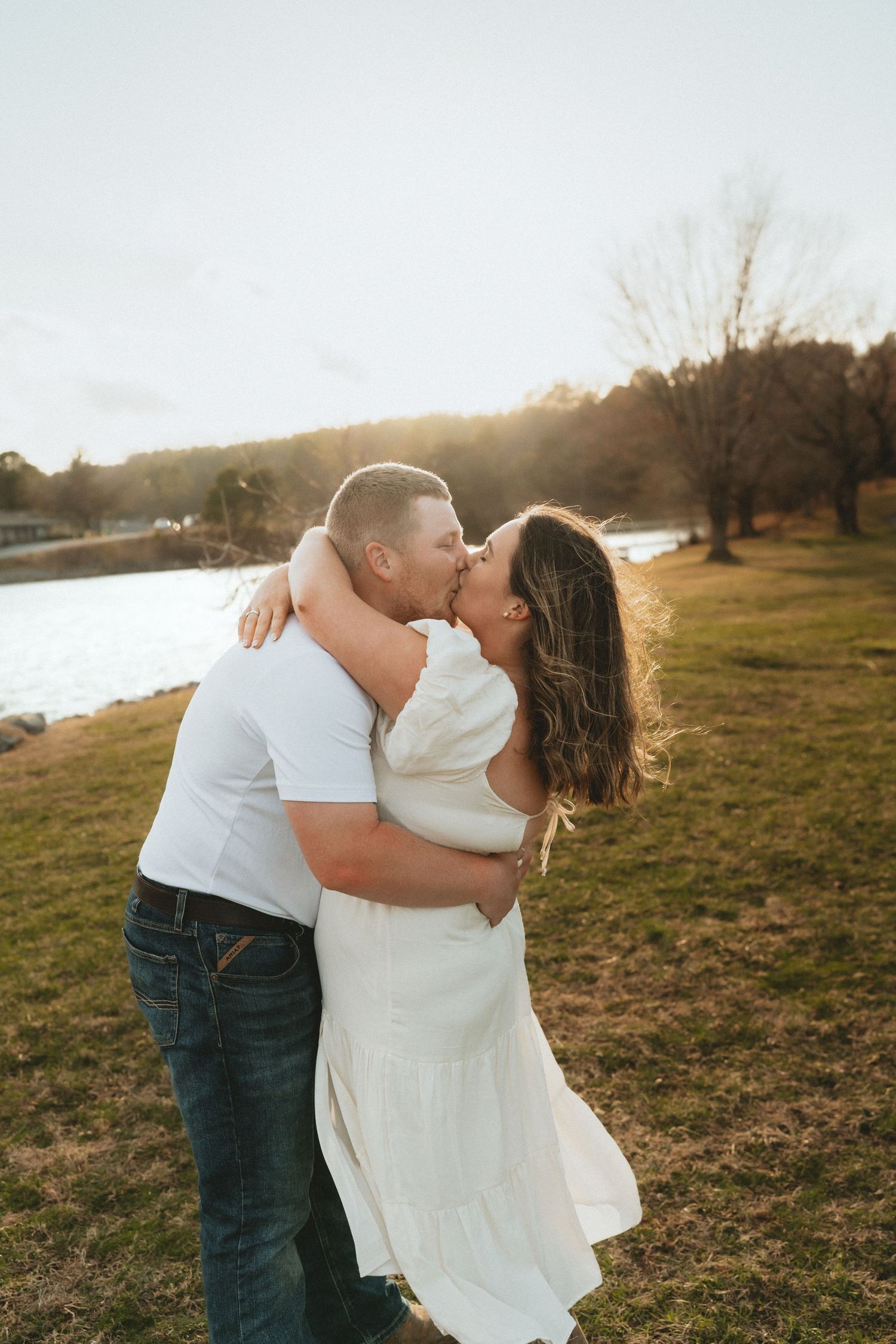 Couple kissing passionately in a grassy field by a lake during a warm golden hour engagement session.