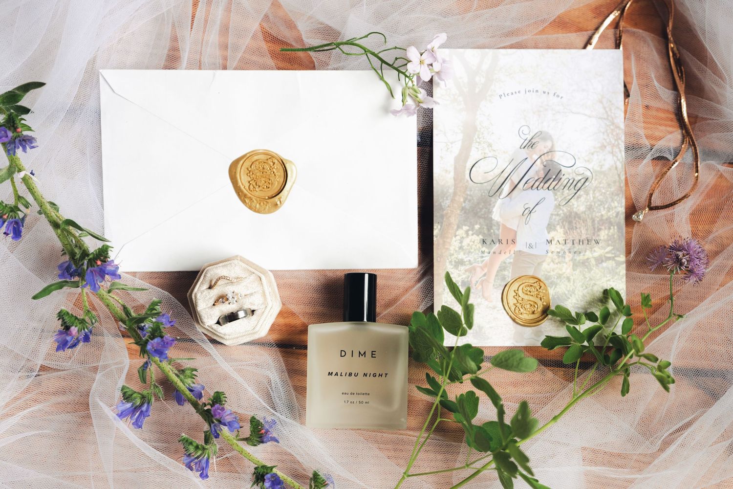 Elegant wedding flat lay featuring gold wax seal envelopes, rings, perfume bottle, and floral accents.