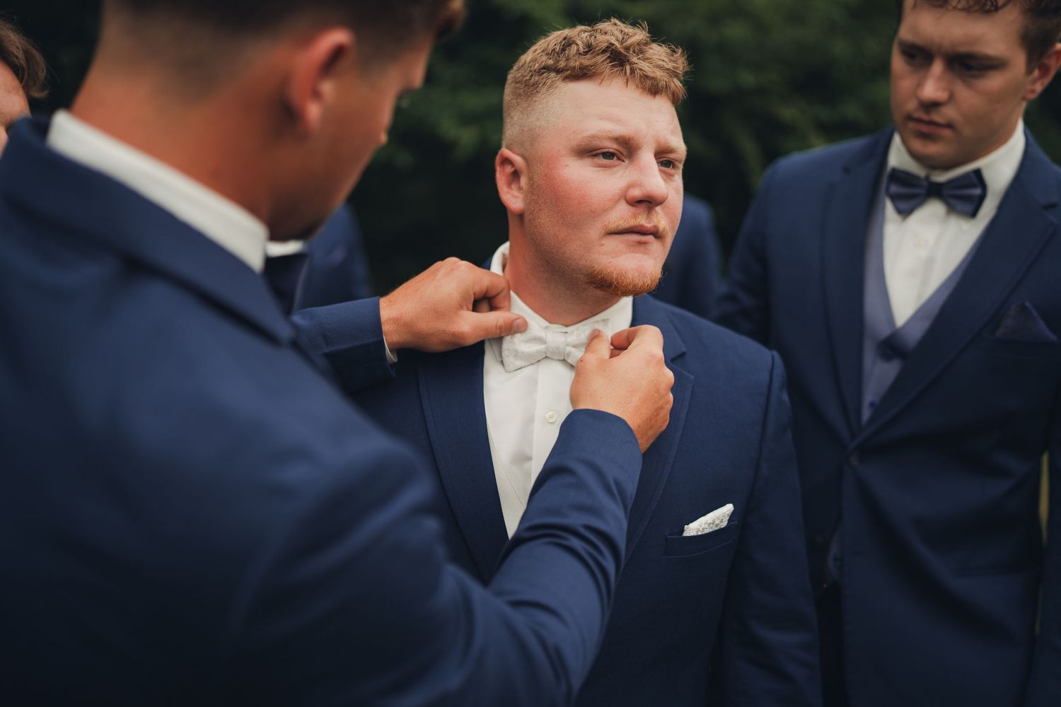 Groomsmen in blue suits adjusting white bow tie on groom before wedding ceremony outdoors.