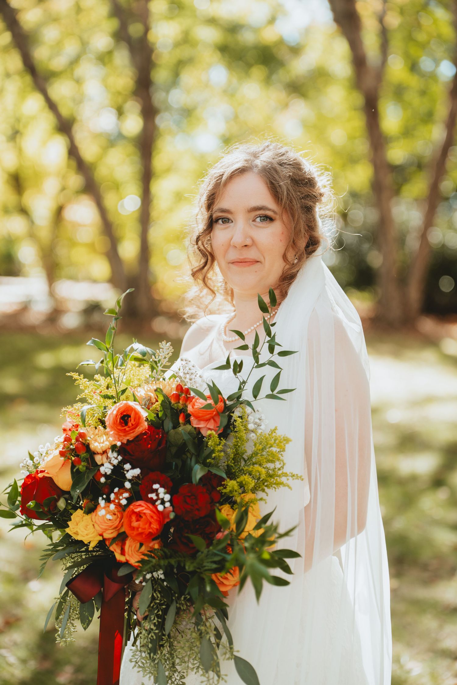 Smiling bride in white gown holds a vibrant bouquet of orange, red, and yellow flowers in a sunny park.