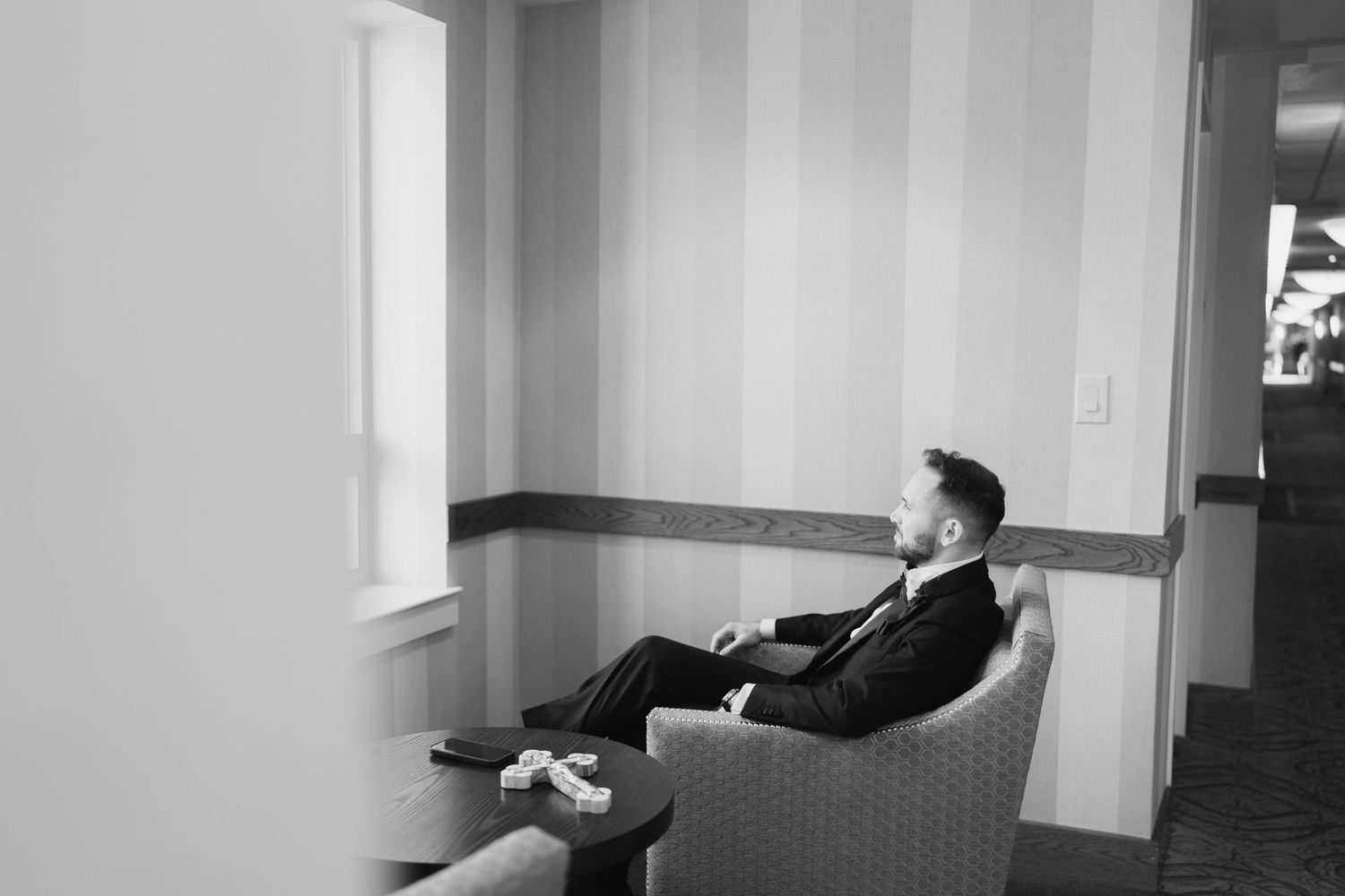 Groom in black suit sitting alone in hotel chair in black and white wedding photo.