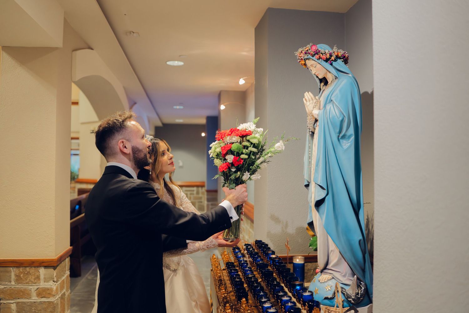 Bride and groom offer floral bouquet to Virgin Mary statue during Catholic church wedding ceremony.