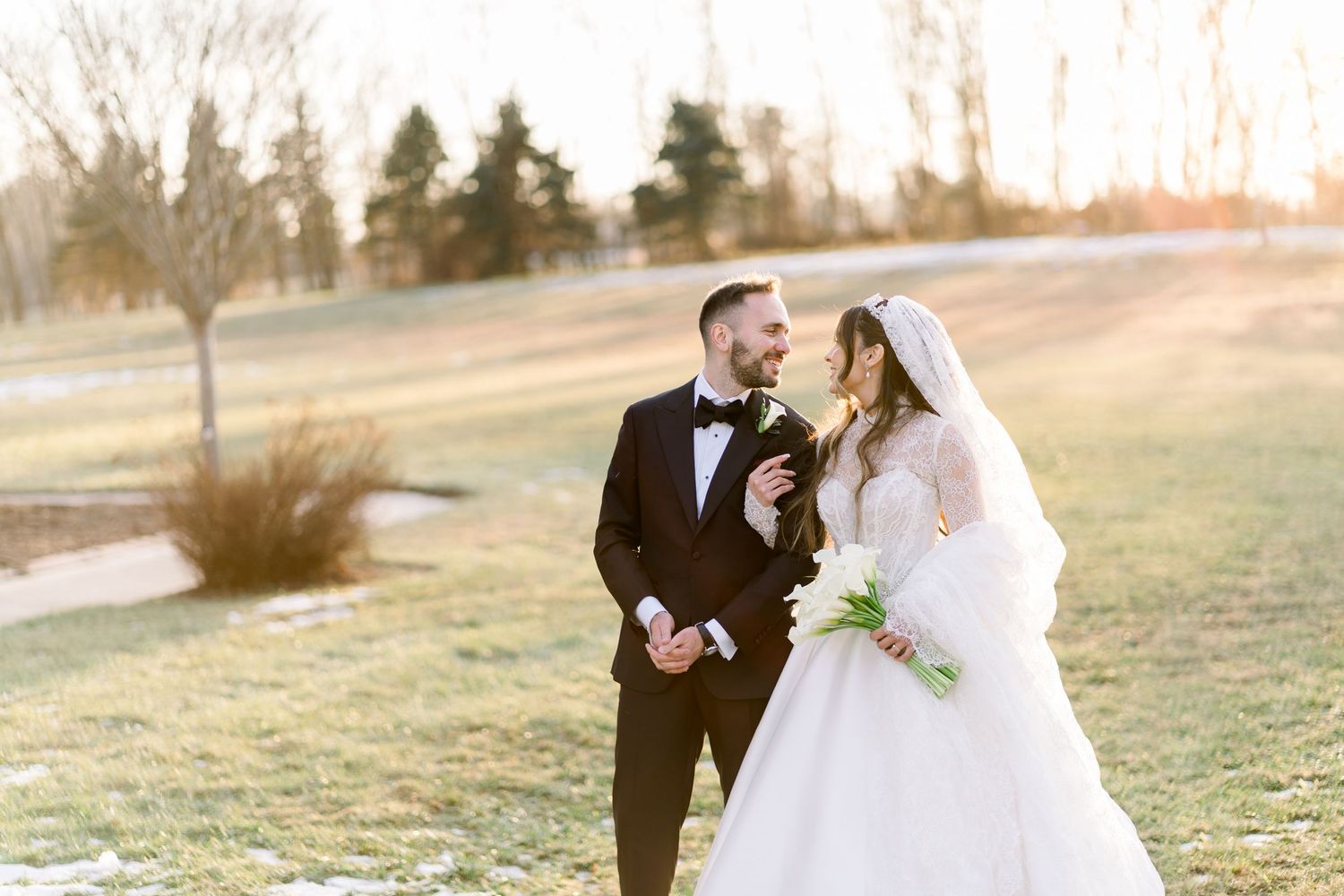 Bride in white lace ball gown and groom in black tuxedo smile at each other on frost-covered lawn at sunset.