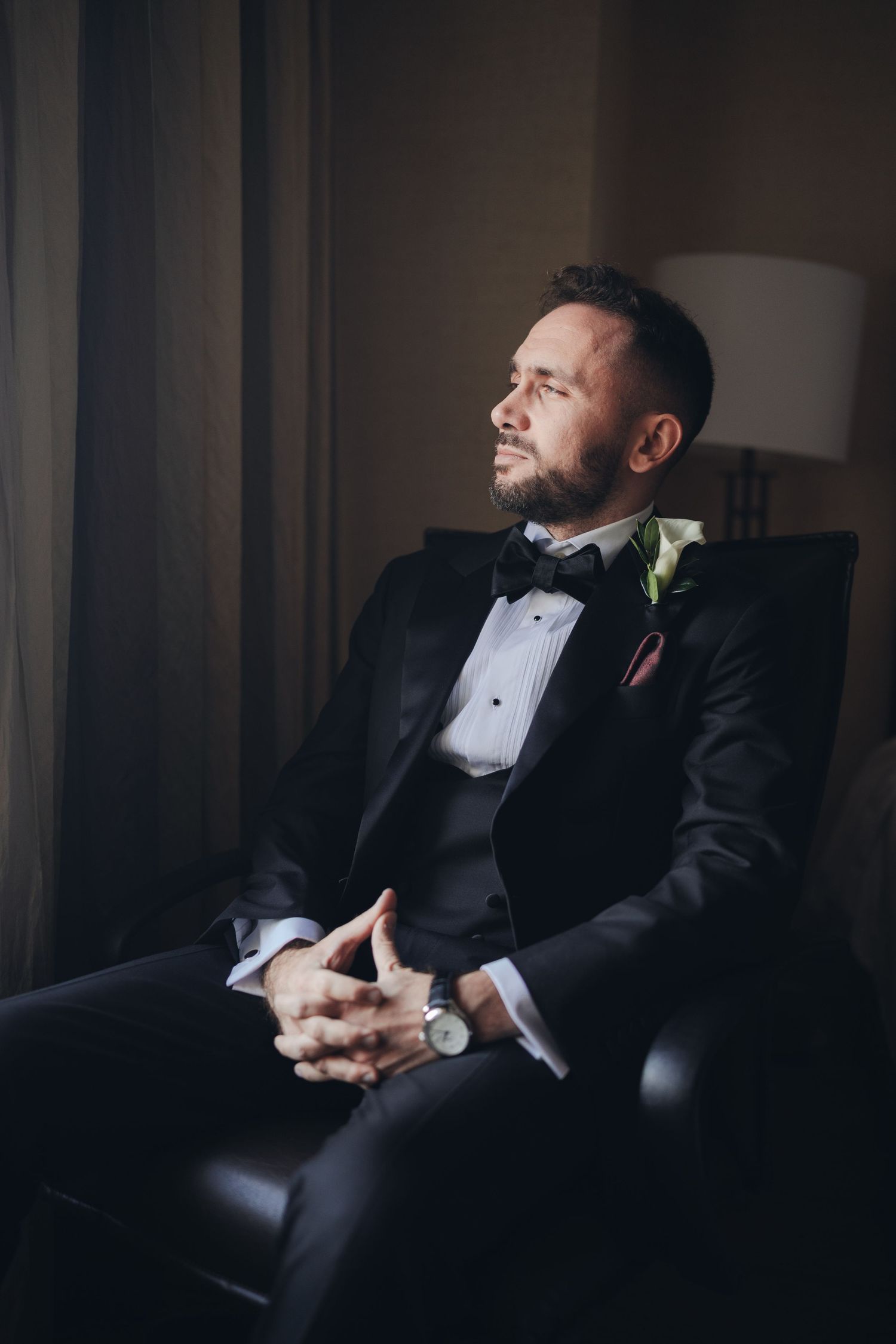 Dapper groom in black tuxedo with bow tie seated in leather chair by a softly lit window.