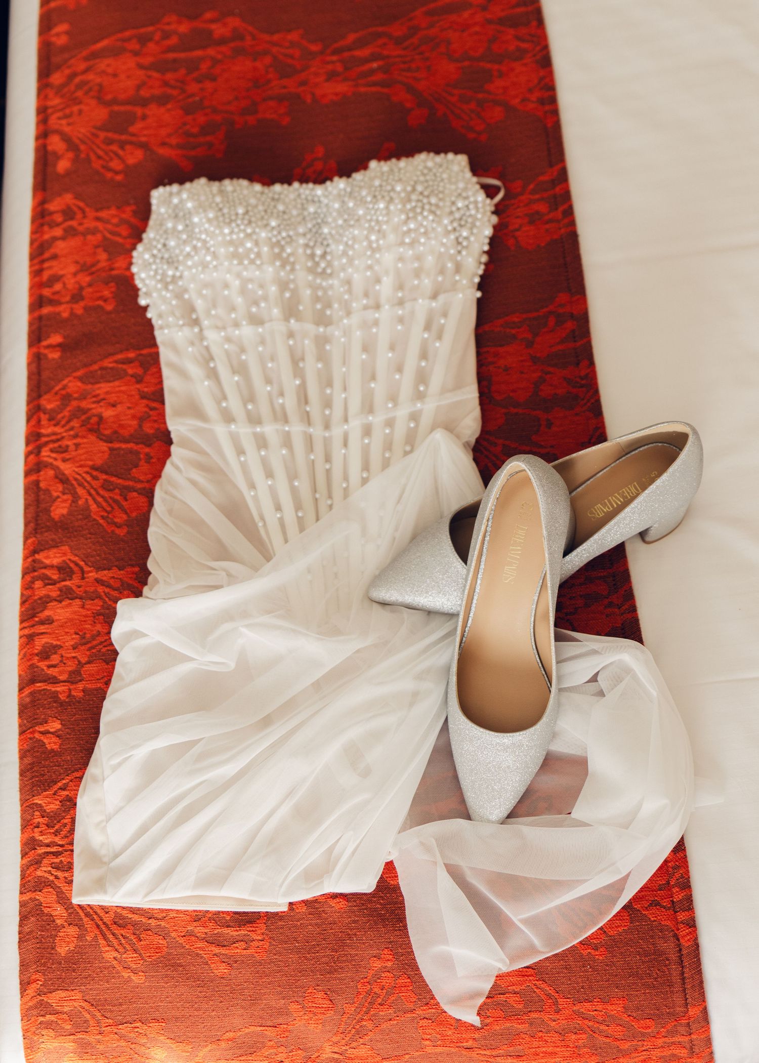 Pearl-embellished strapless wedding dress with silver glitter heels on red floral fabric.