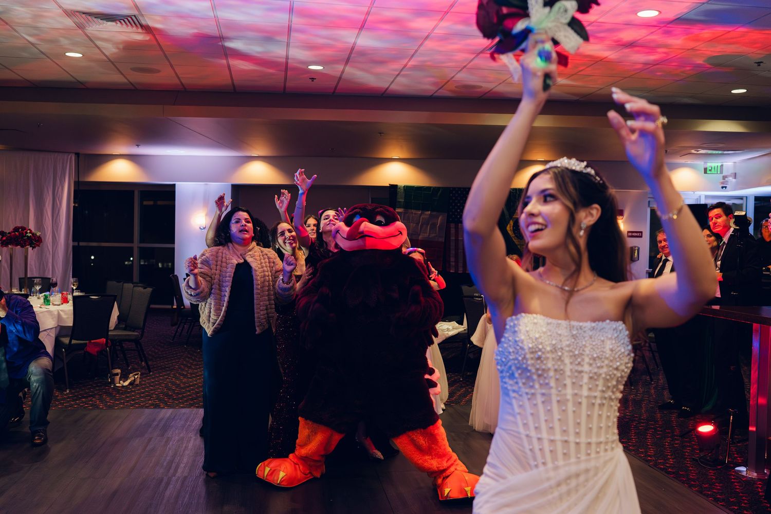 Bride in strapless white gown raises bouquet while dancing with mascot character at vibrant wedding reception.