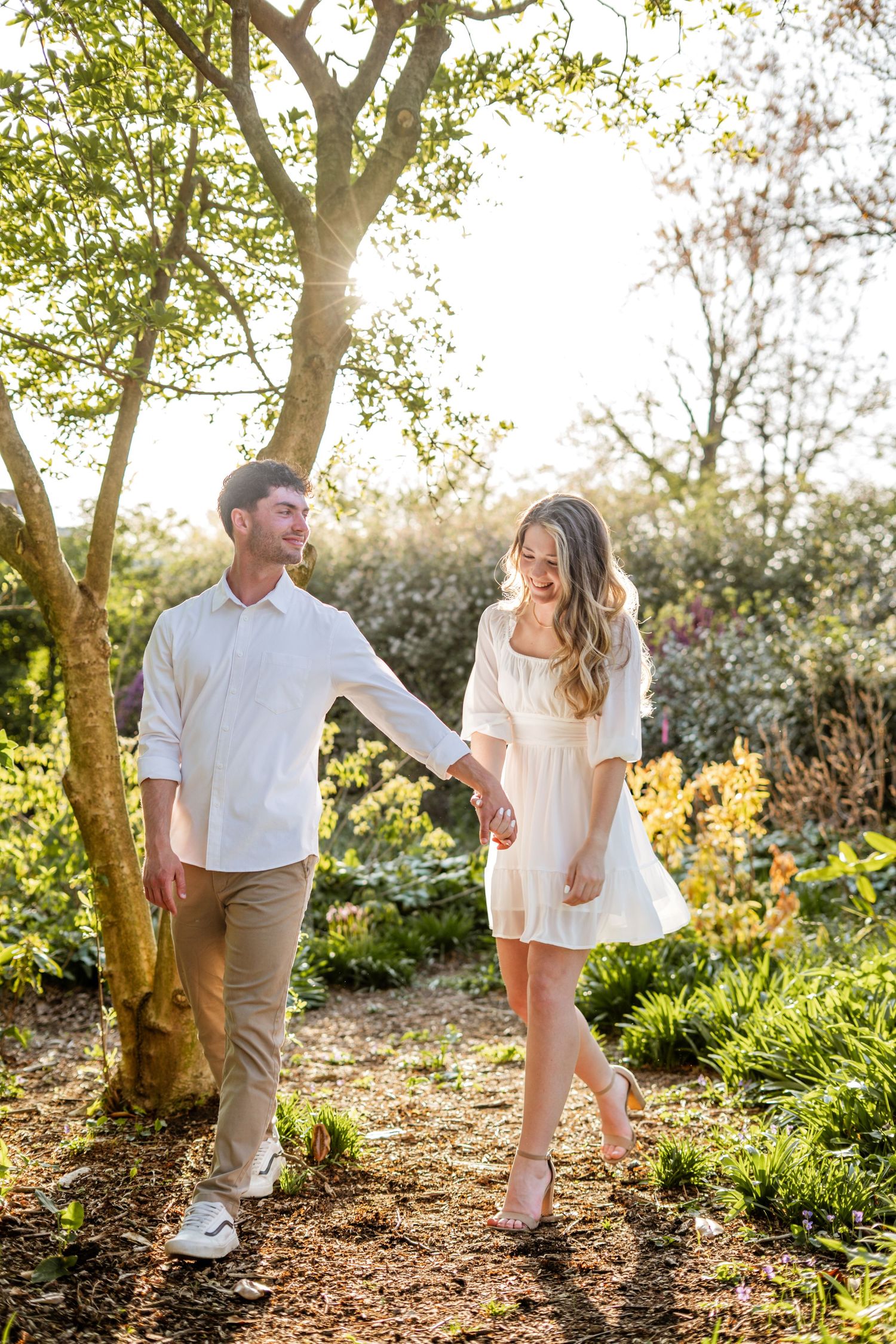 Couple in white outfits holding hands and smiling while walking through a sunlit garden in spring.