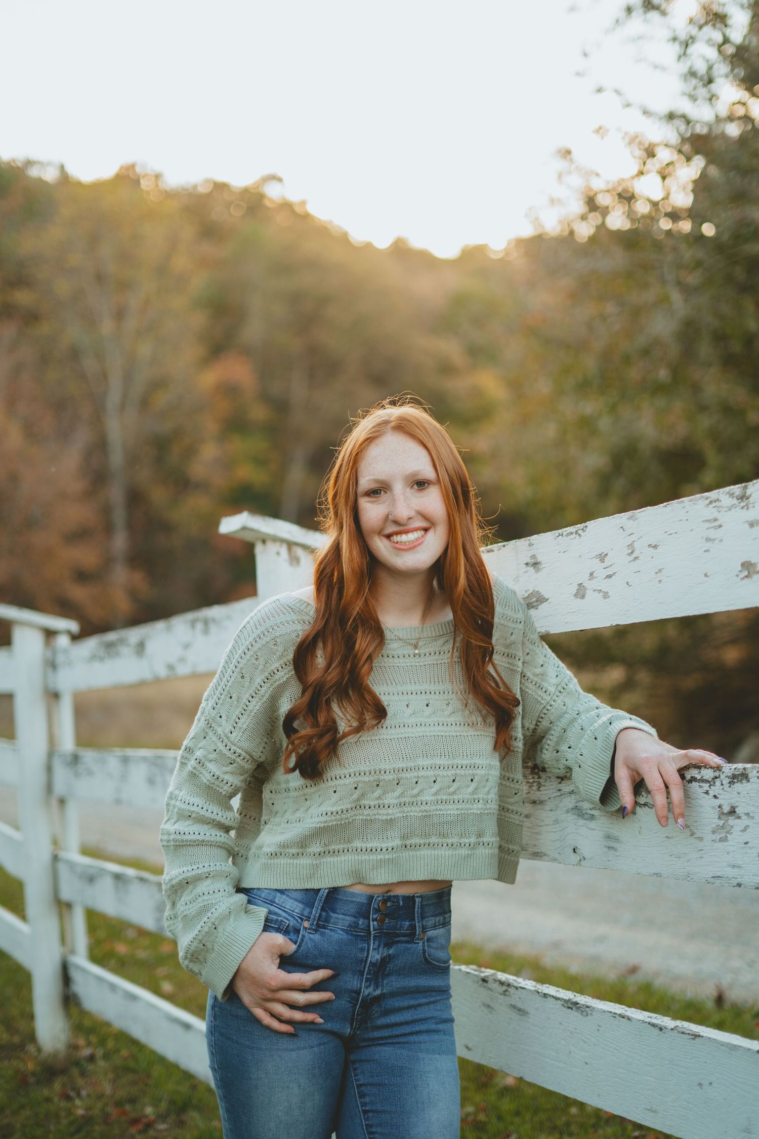 Smiling redhead girl in mint sweater leaning on white fence in autumn landscape.