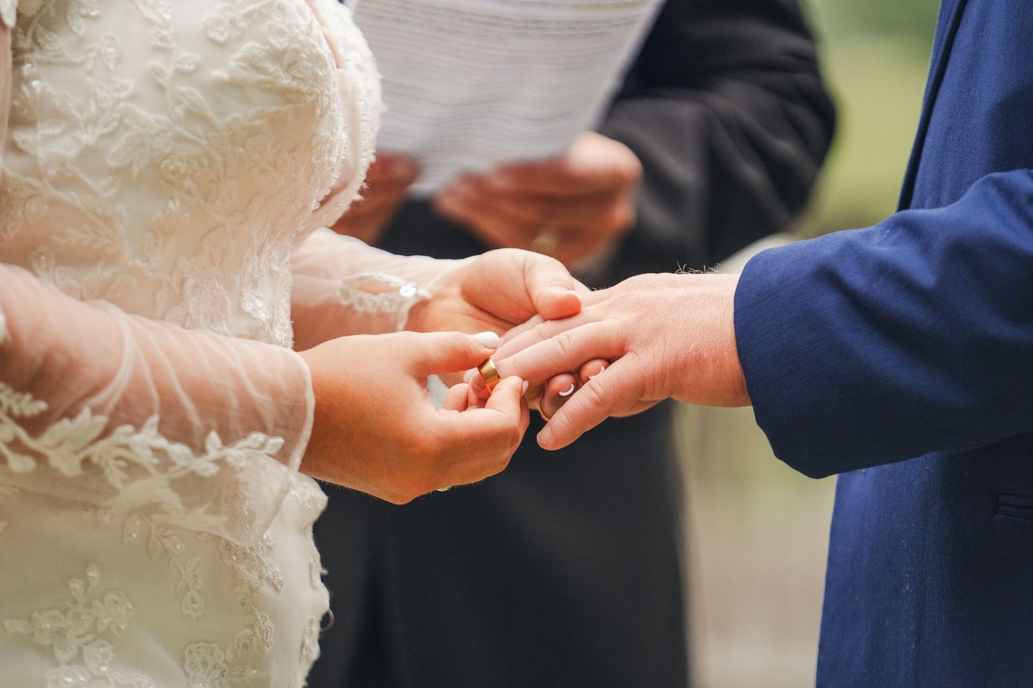 Bride placing a wedding ring on groom's finger during an outdoor ceremony, with officiant holding vows in background.