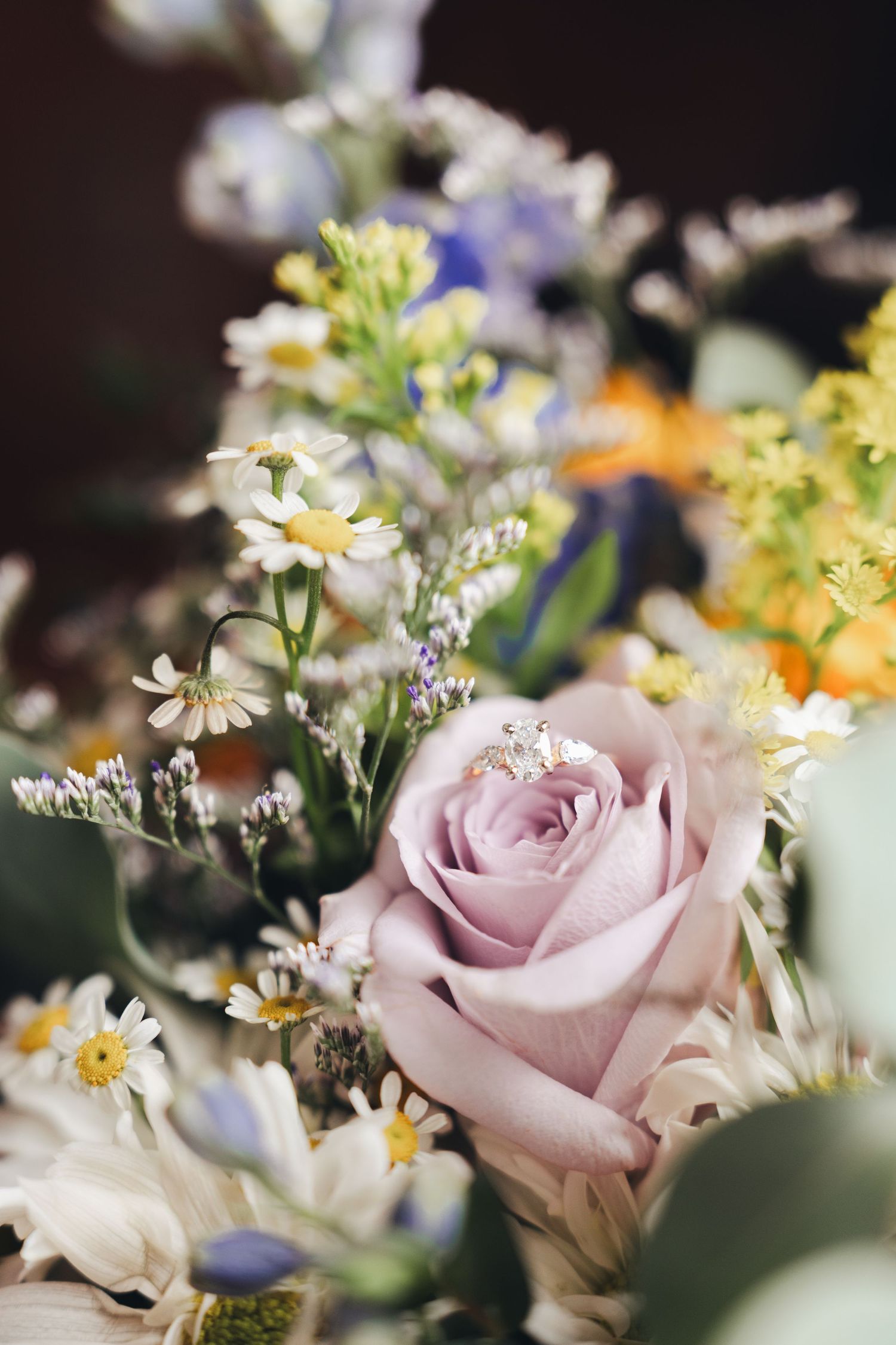 A diamond engagement ring resting on a soft pink rose in a colorful wildflower bouquet.