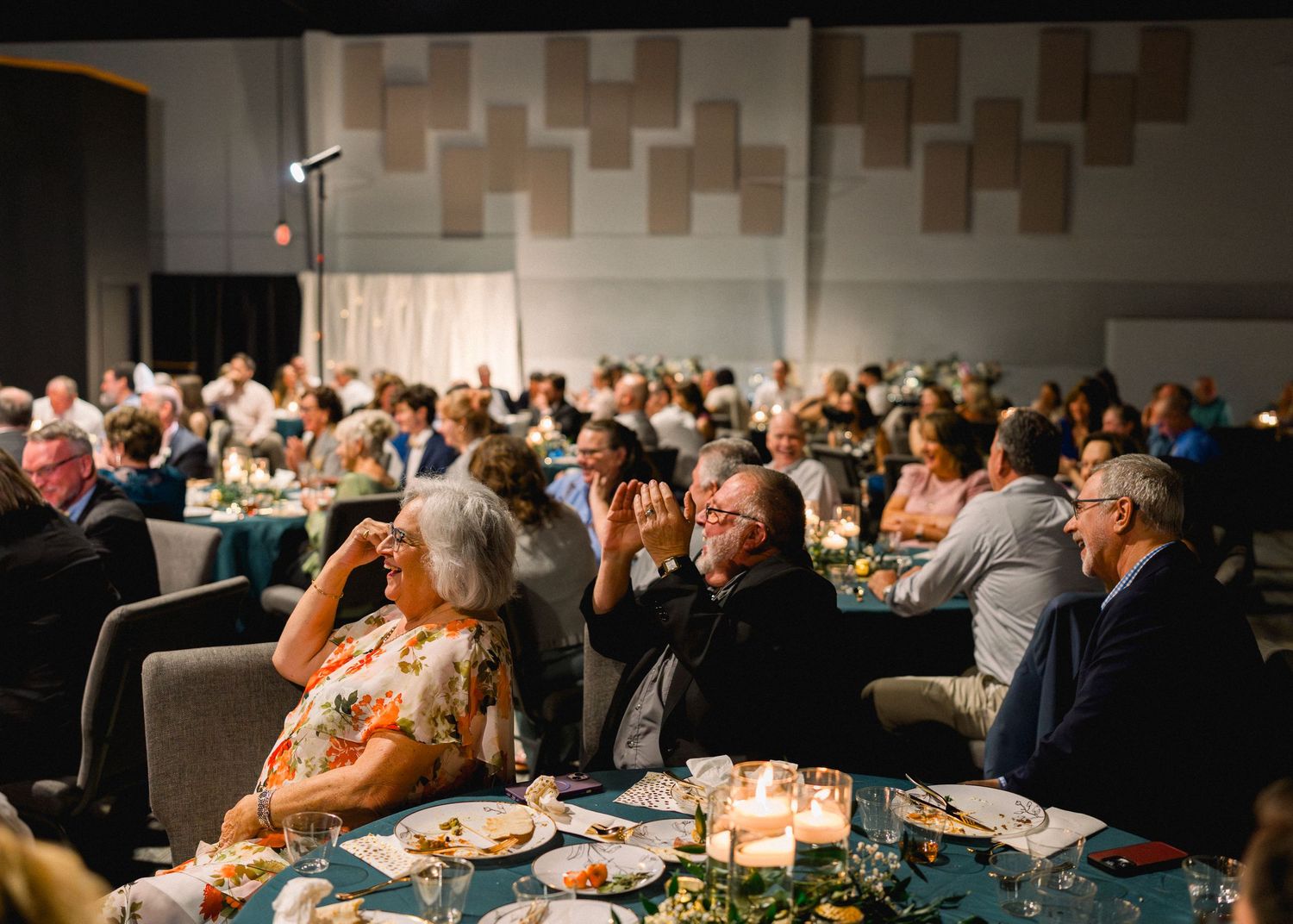 Large wedding reception hall filled with guests seated at round tables with candlelit centerpieces and teal linens.
