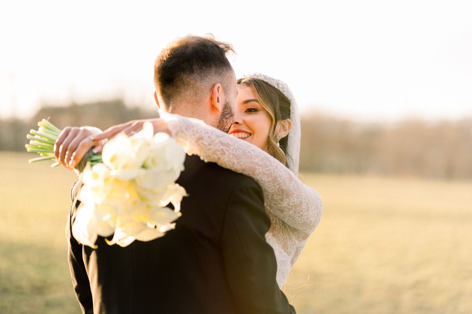 Radiant bride in lace gown embraces groom at sunset, holding white bouquet in golden field.