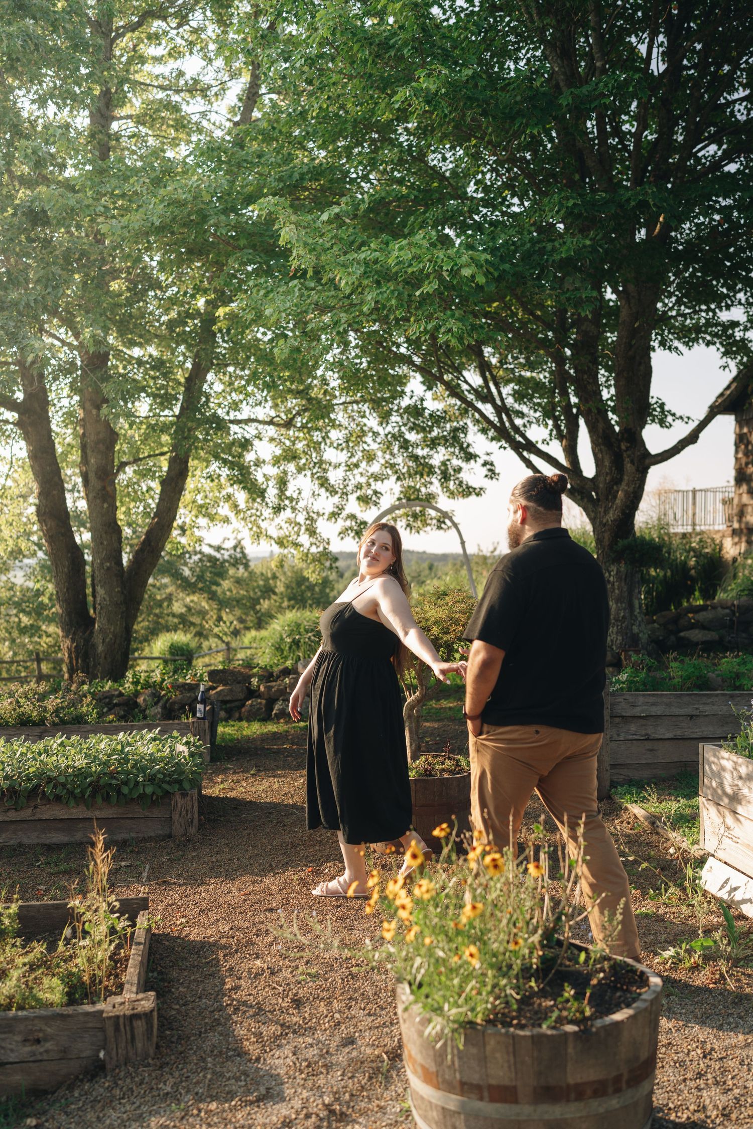 Couple holding hands and laughing together in a sunlit garden with raised flower beds and trees.