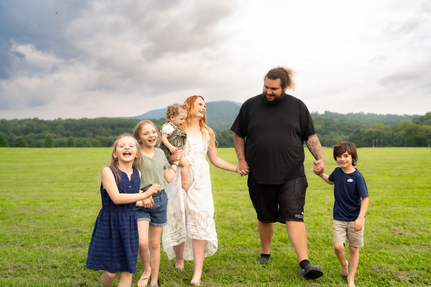 Happy family of five walking together across a lush green field with mountain views behind them.
