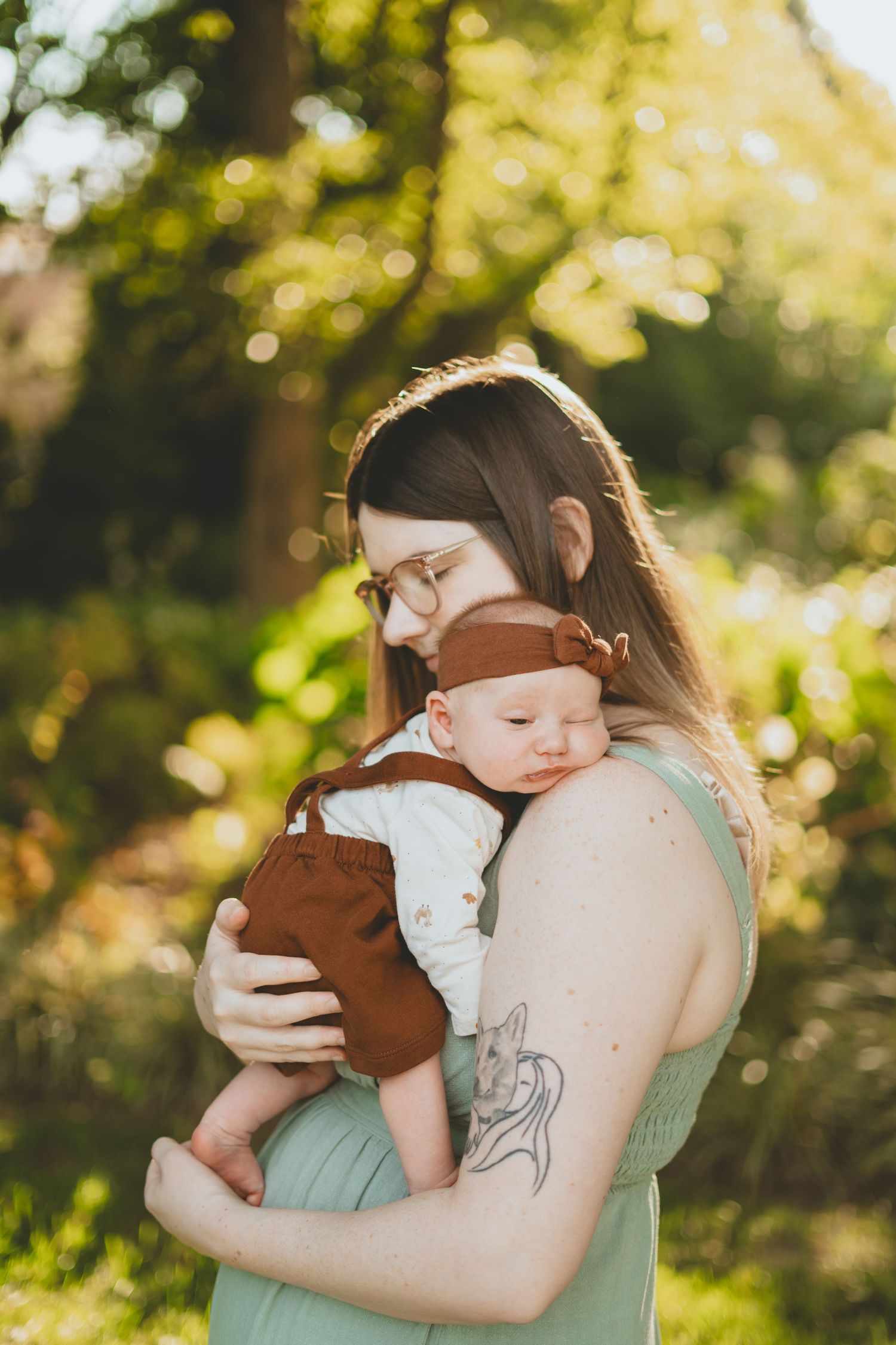 Mother gently holds sleeping newborn baby in brown outfit in warm sunlit garden setting.