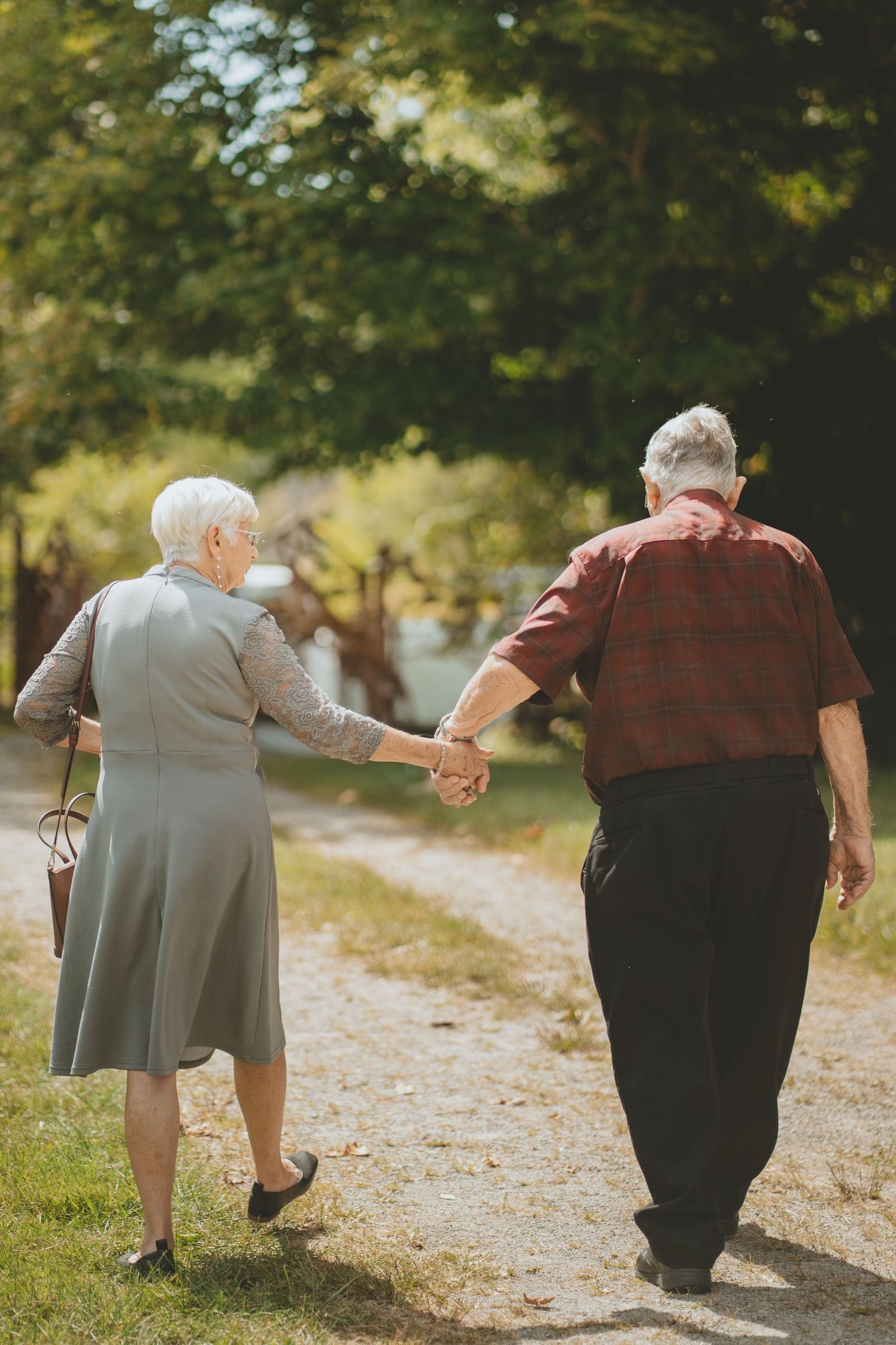 Elderly couple holding hands while walking along a sunny garden path surrounded by green trees.