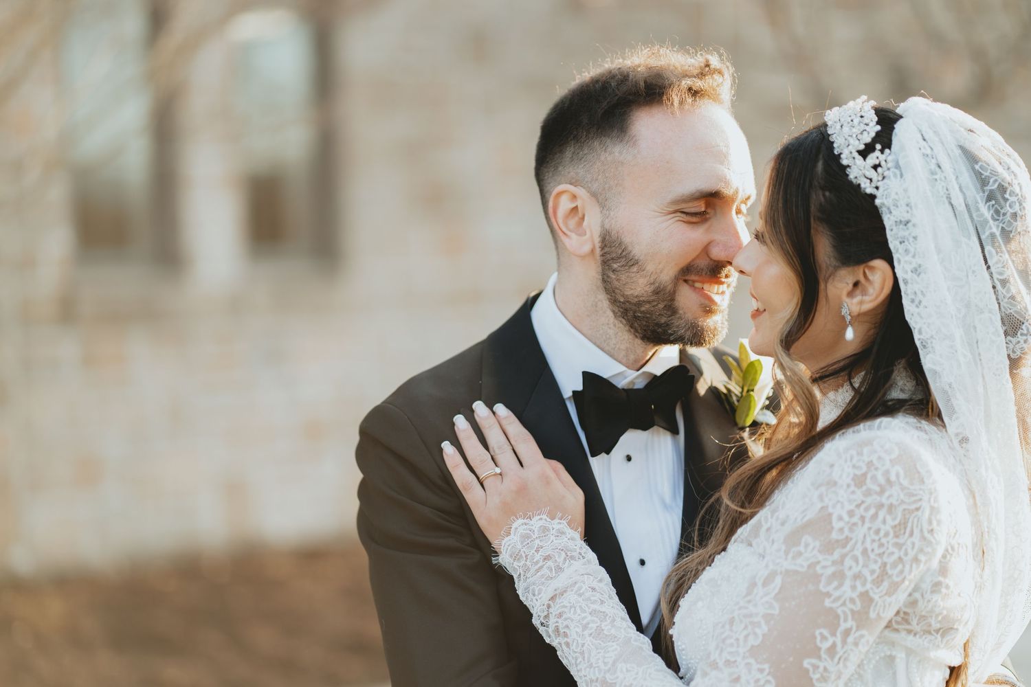 Bride and groom share a romantic moment, smiling closely together, bride wearing lace veil and groom in black tuxedo.