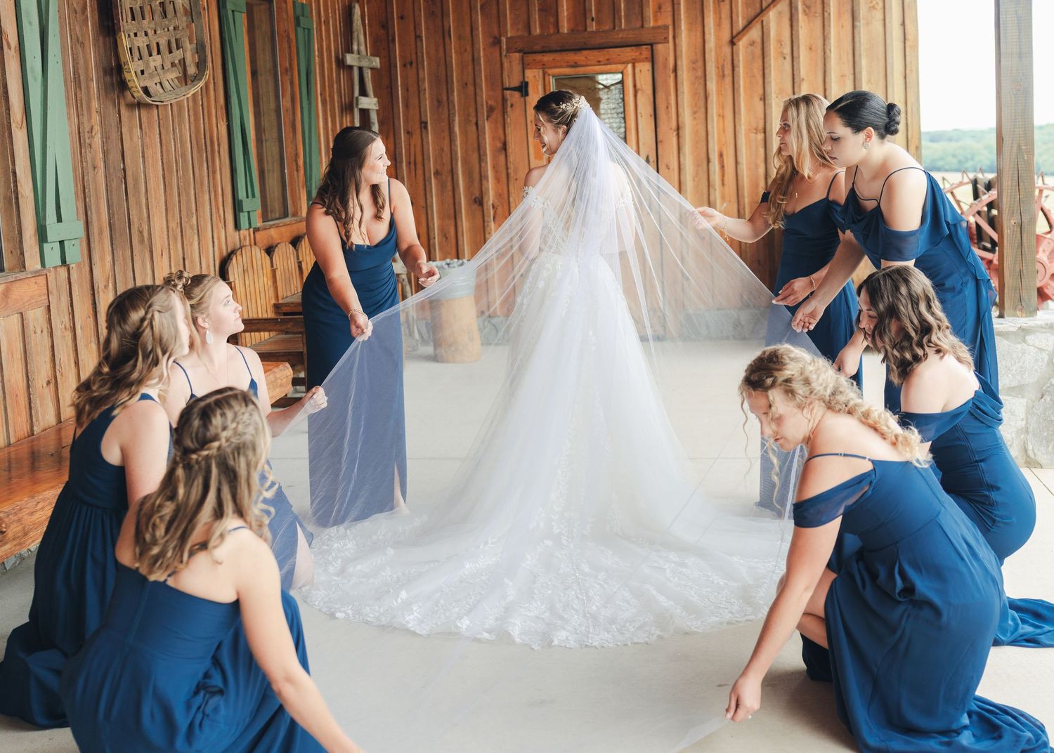Bridesmaids in navy blue dresses arrange a bride's white lace veil in a rustic barn wedding venue.