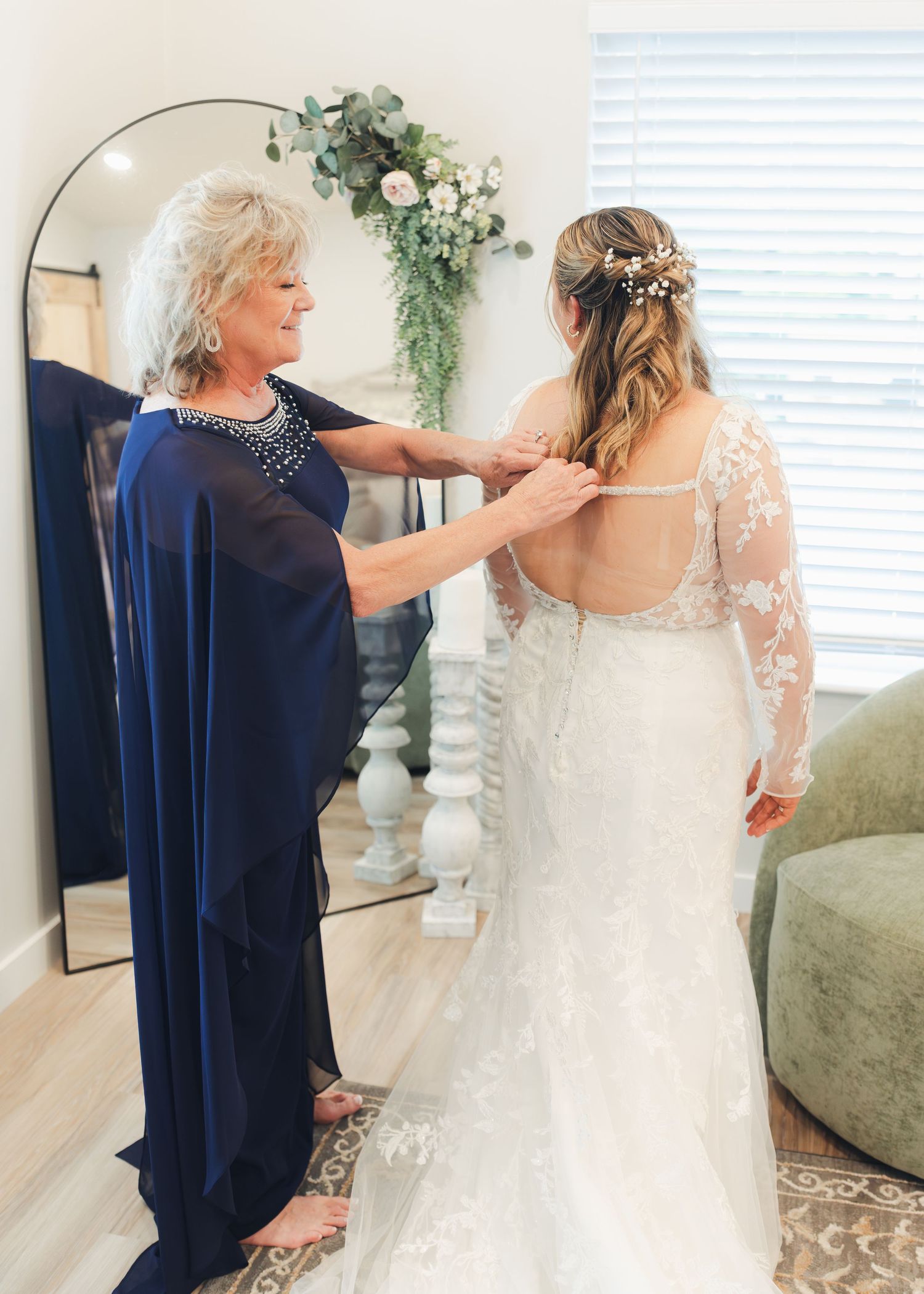 Mother helping bride zip up her white lace wedding gown in a bright, elegant bridal suite.