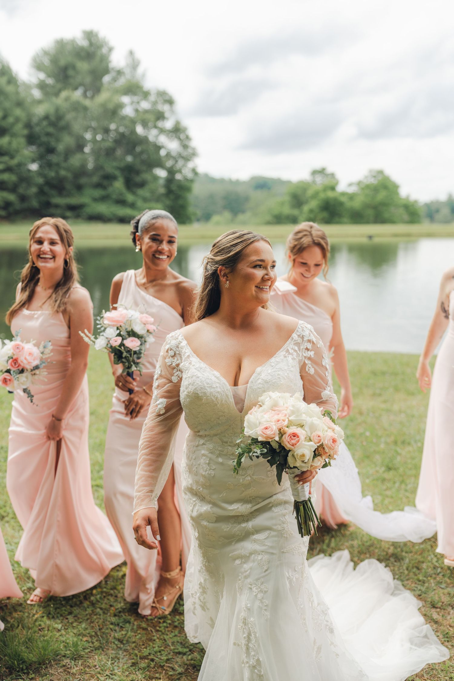 Bride in lace gown walks with bridesmaids in blush pink dresses carrying bouquets near a scenic pond.