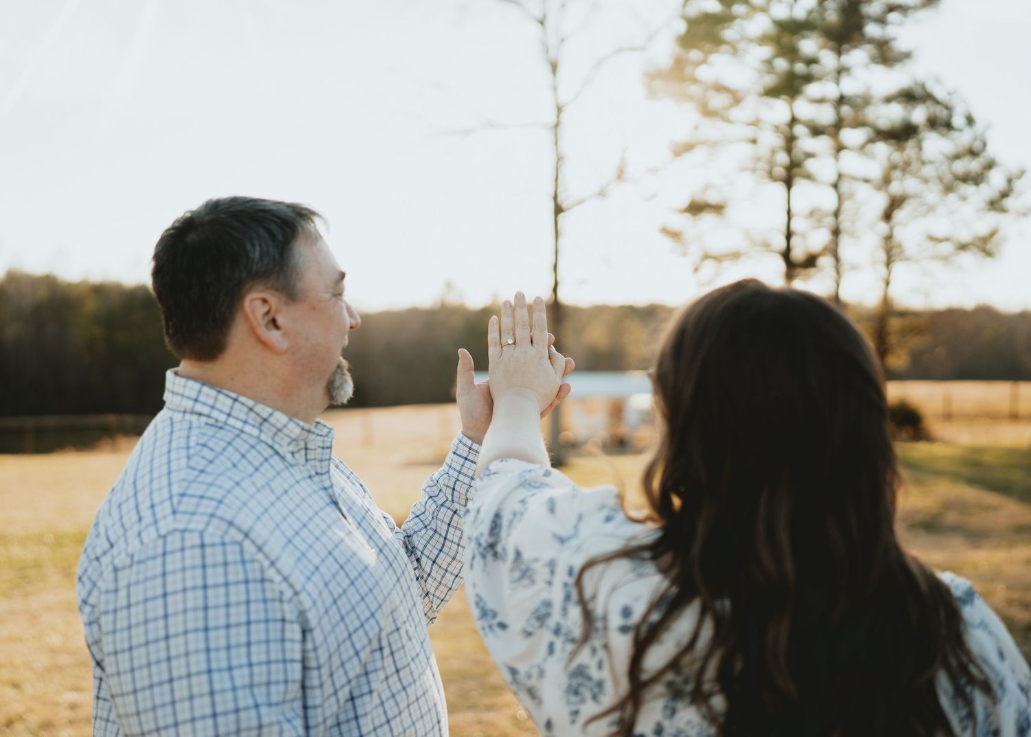 Engaged couple holding up her diamond ring together in a golden hour outdoor farm setting.