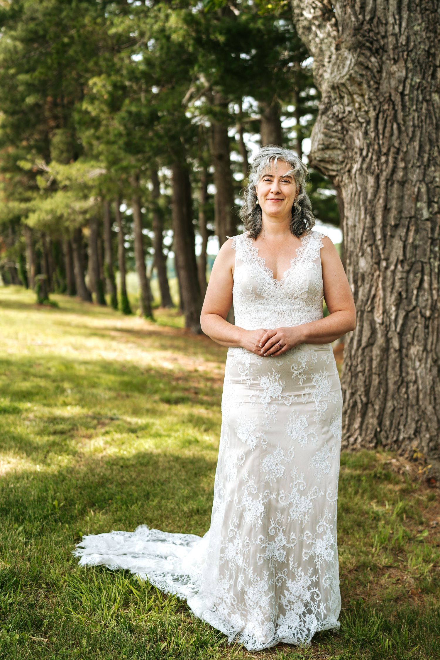 Elegant older bride in lace wedding dress with train posing beside tall pine tree outdoors.