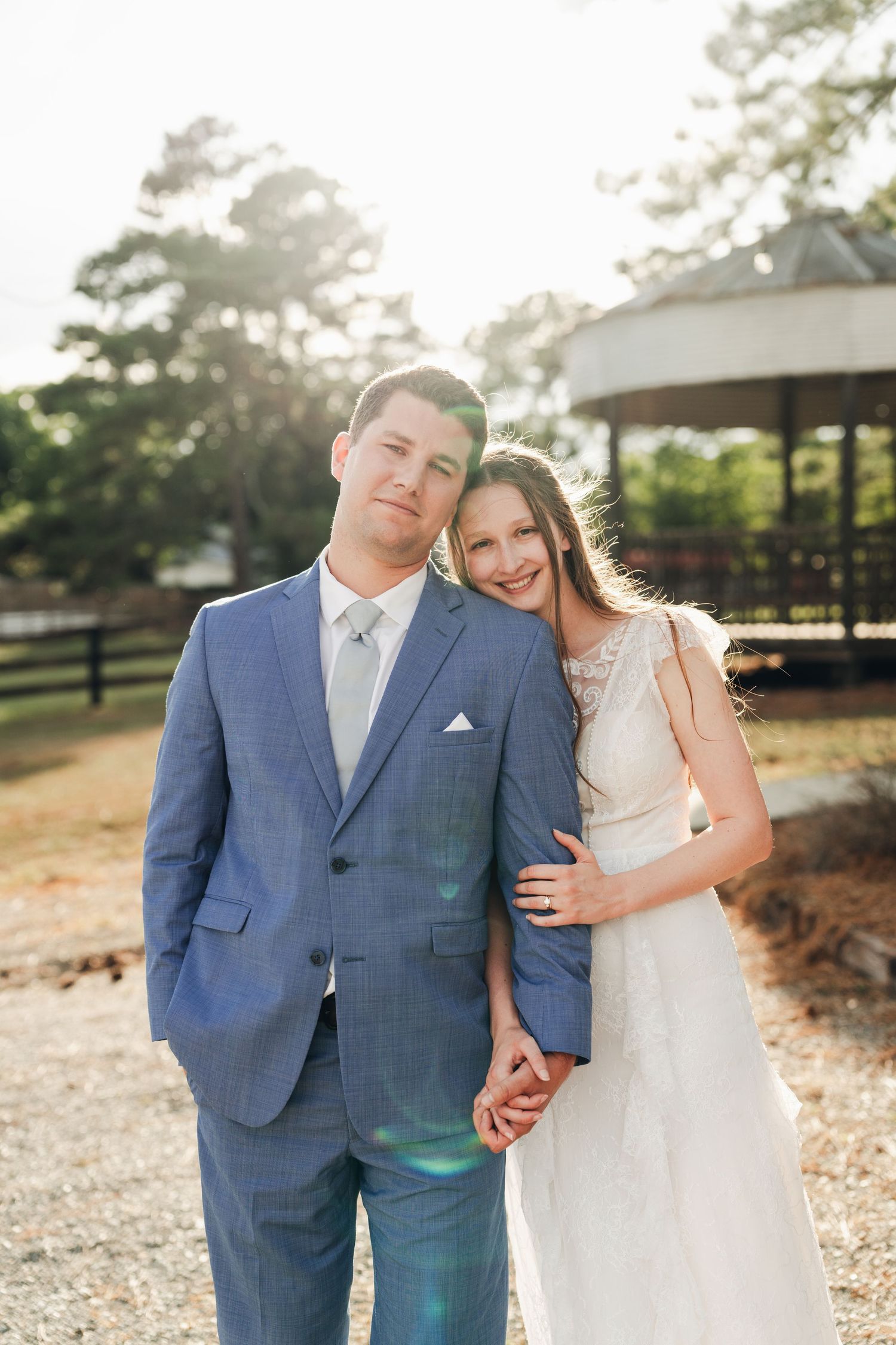 Bride and groom posing together in golden sunlight near a rustic gazebo at outdoor venue.