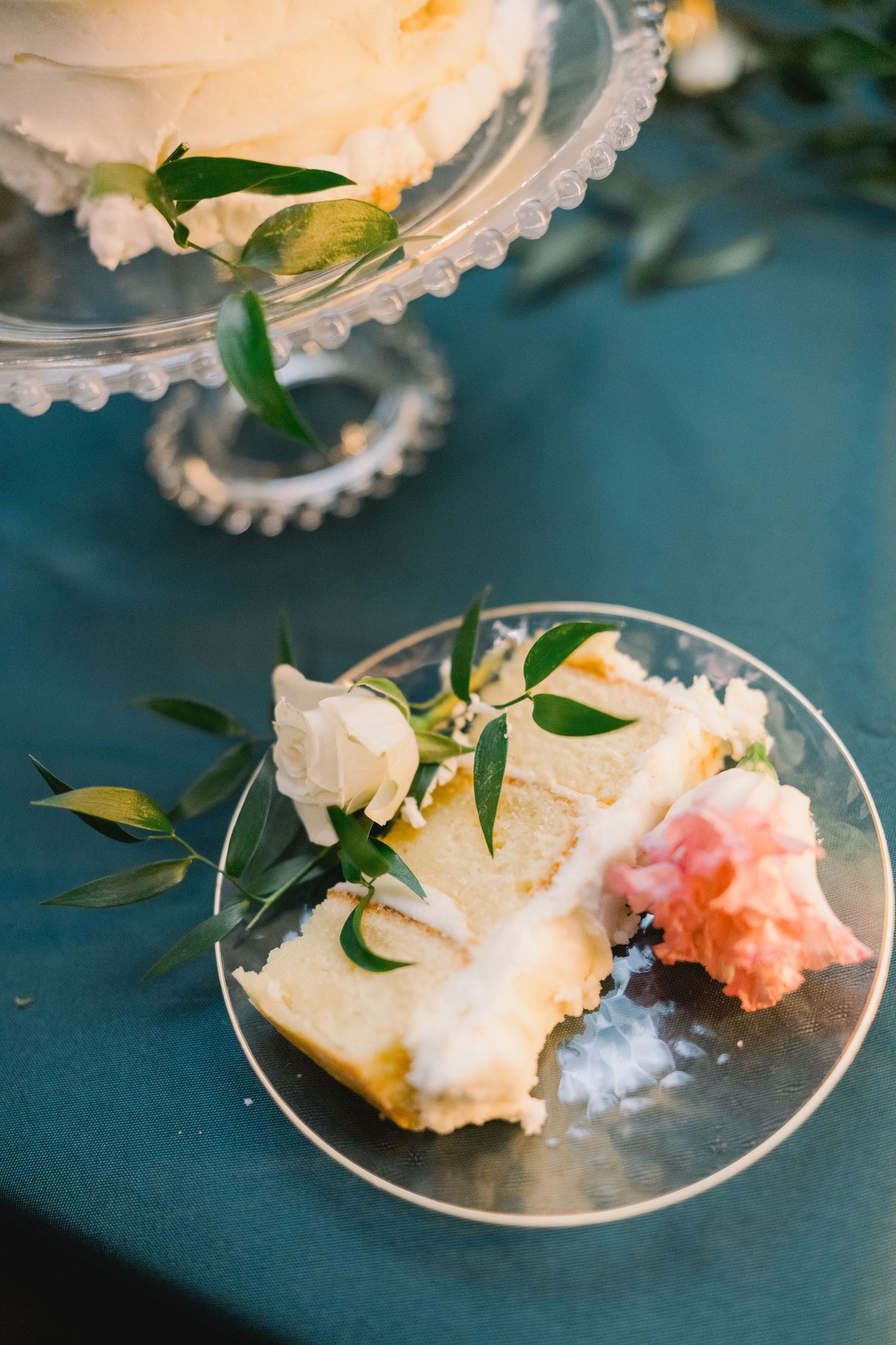 Slice of white wedding cake with cream frosting and white rose garnish on glass plate.