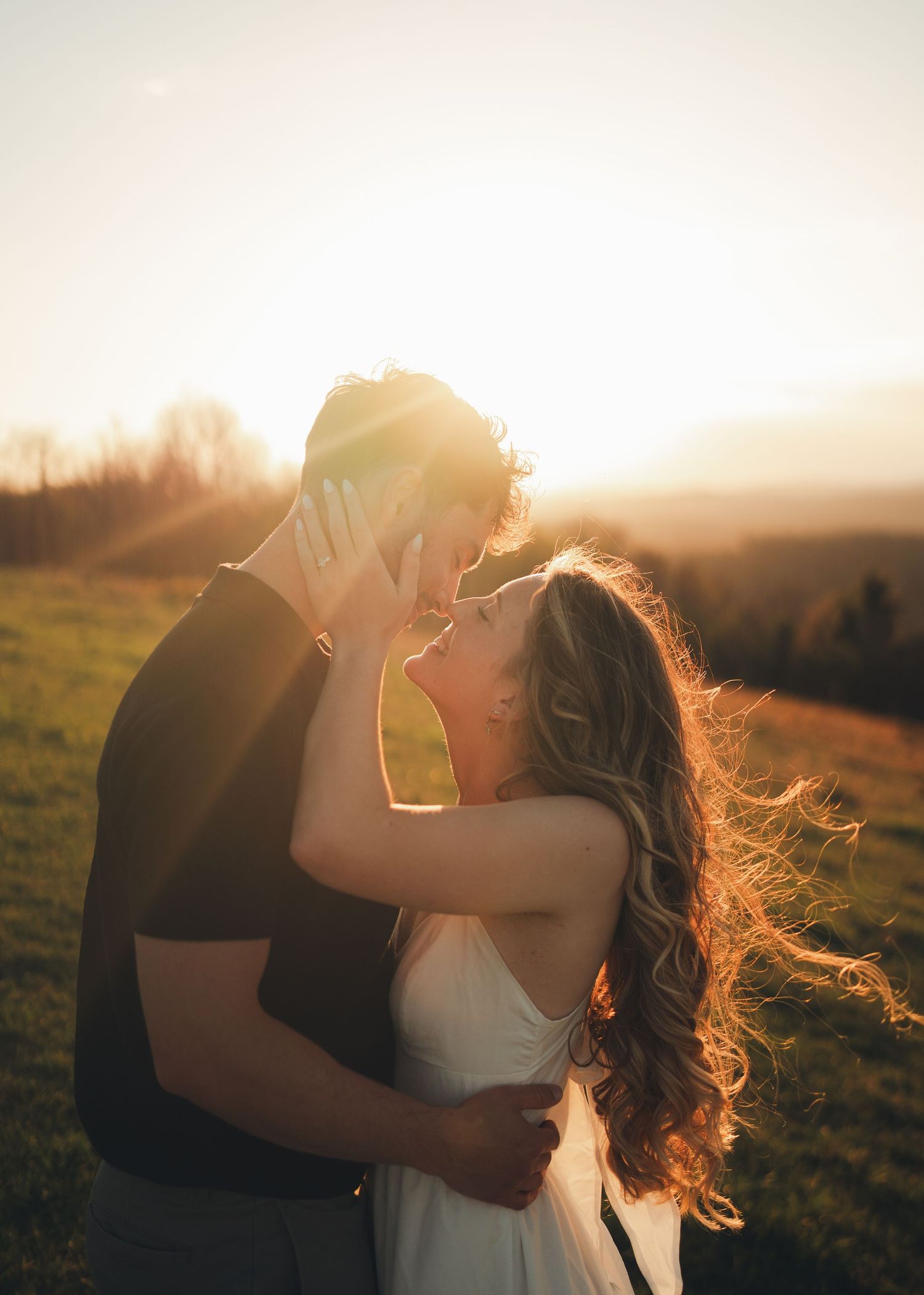 Couple sharing a romantic almost-kiss at golden sunset on a scenic green hillside, woman in white dress with flowing hair.
