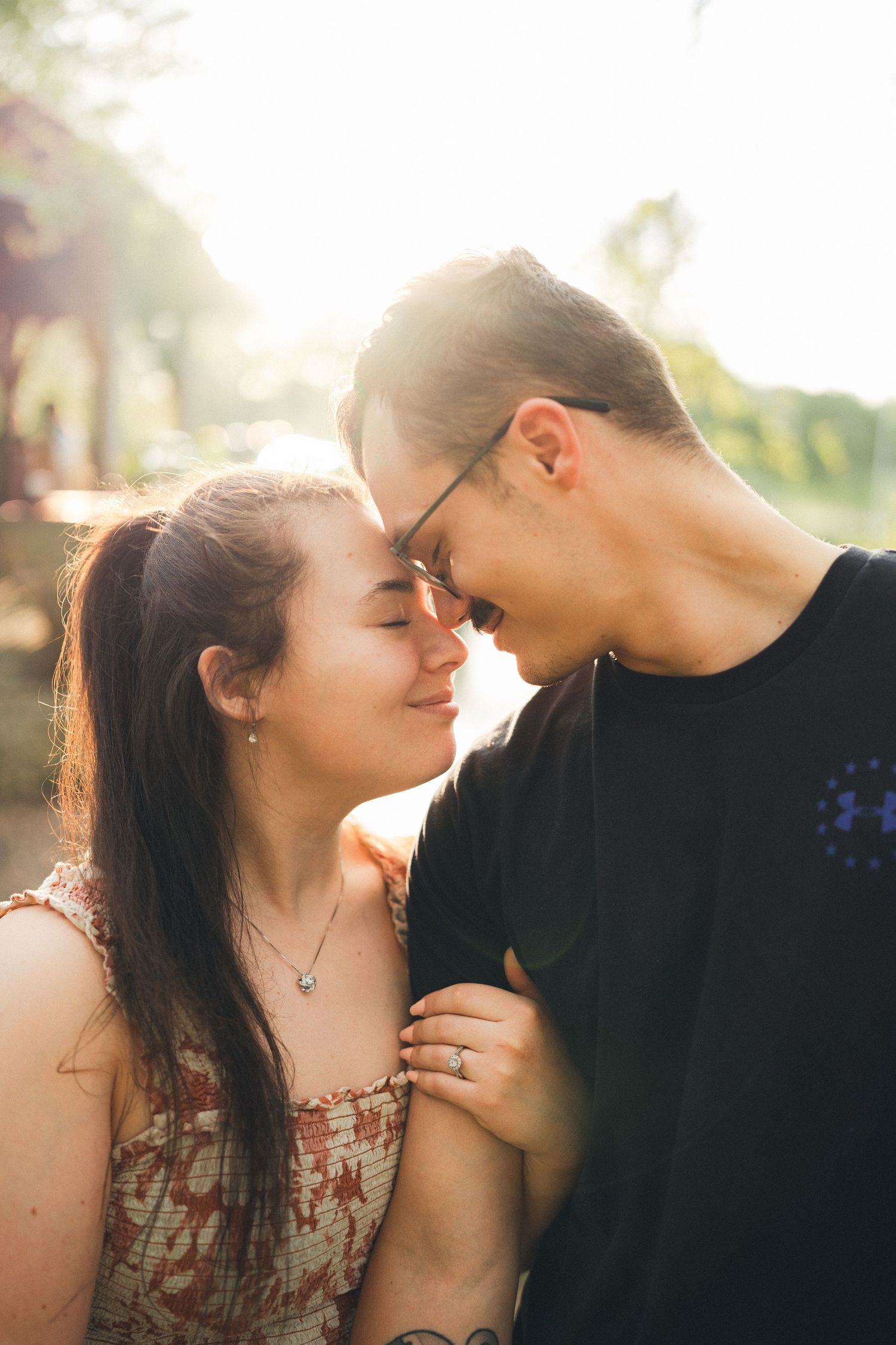 Couple shares intimate forehead touch in warm sunlit park, smiling tenderly at each other.