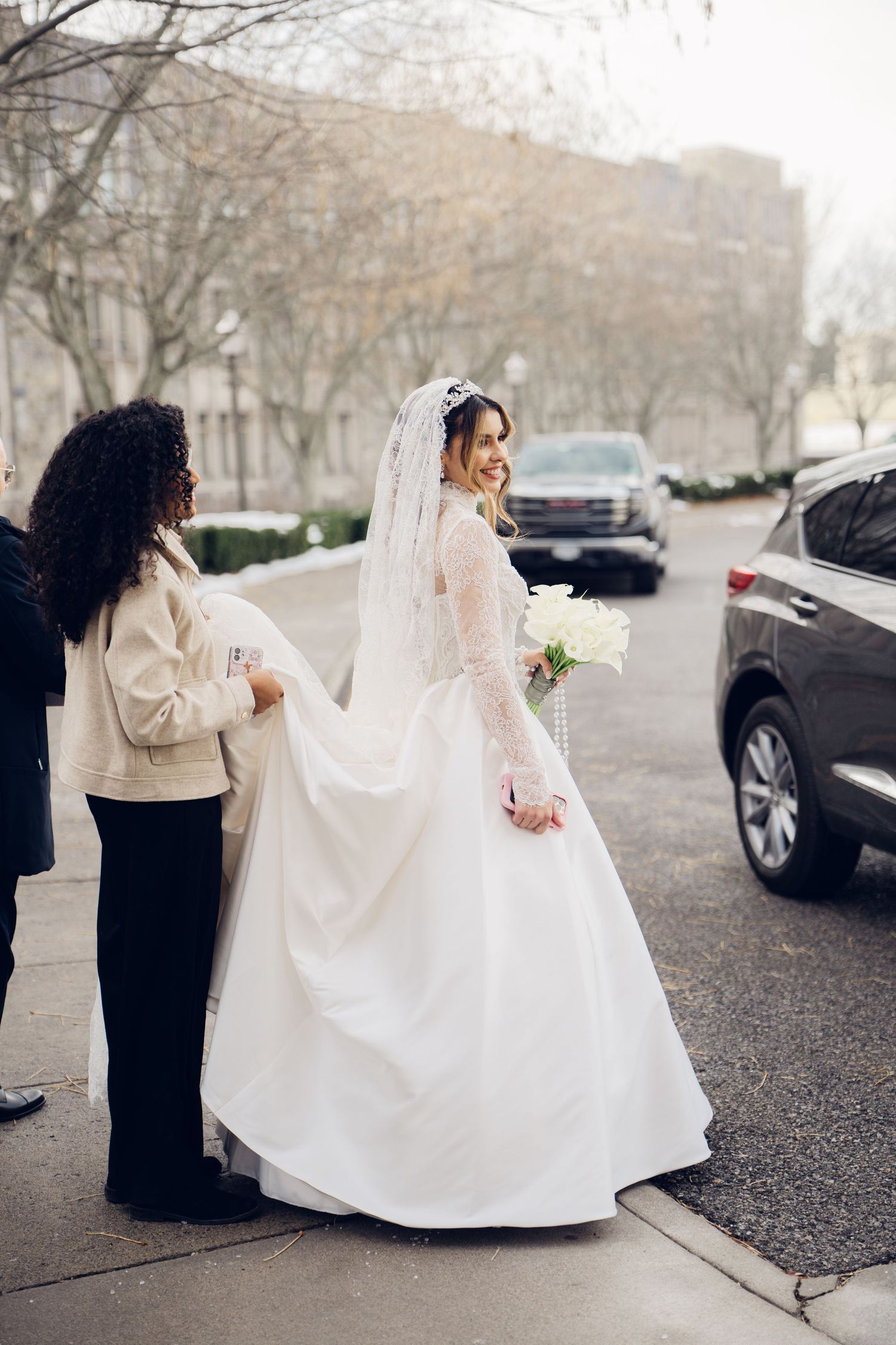 Smiling bride in white lace gown and veil holding bouquet while bridesmaid adjusts train outdoors.