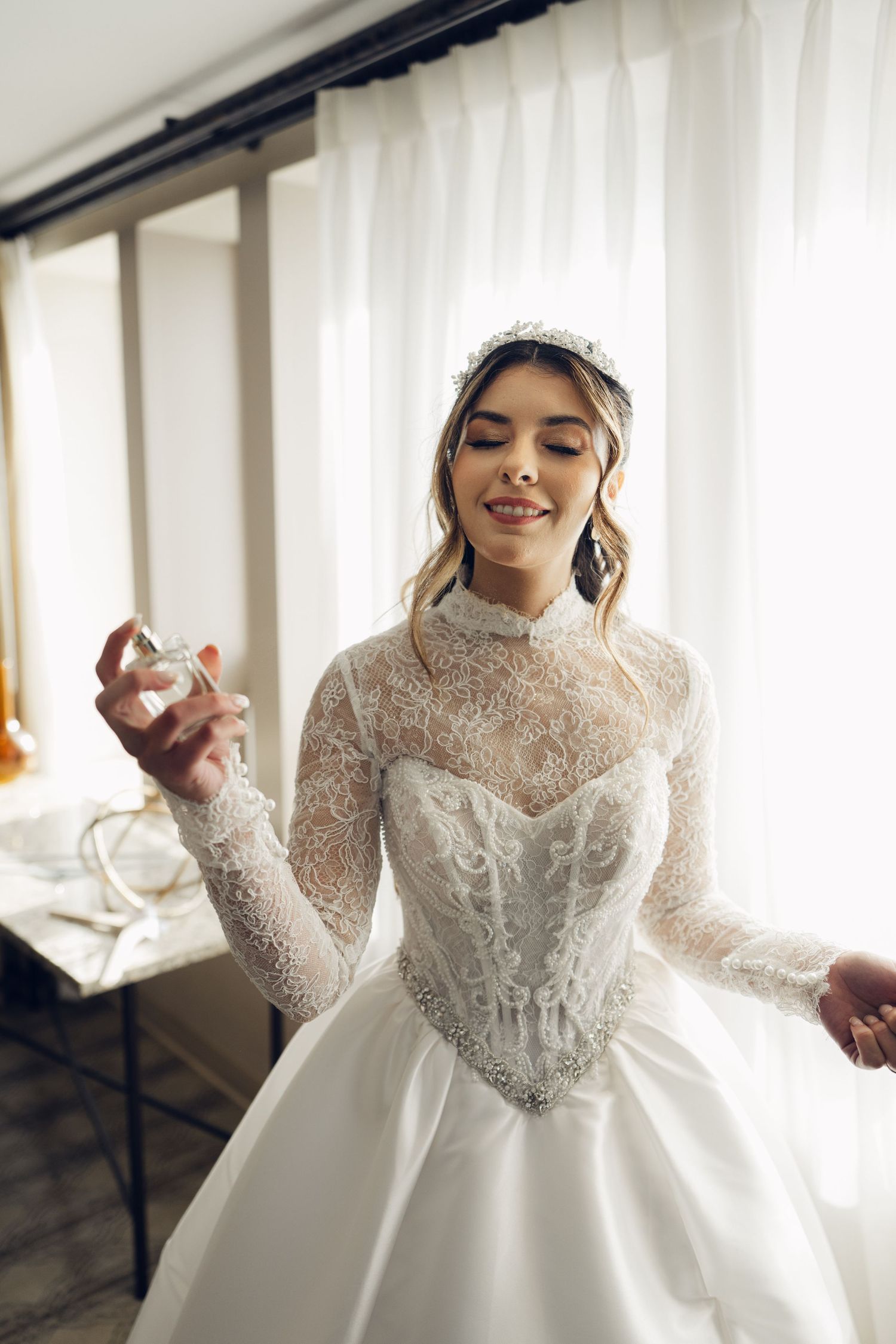Radiant bride in lace high-neck wedding gown and crystal tiara applying perfume before ceremony.