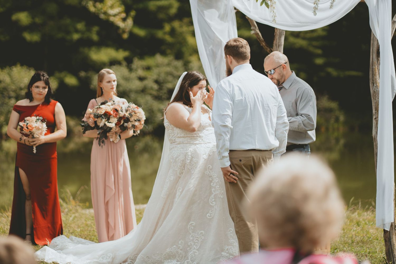 Bride wipes tears during emotional outdoor wedding ceremony with bridesmaids in red and pink gowns.
