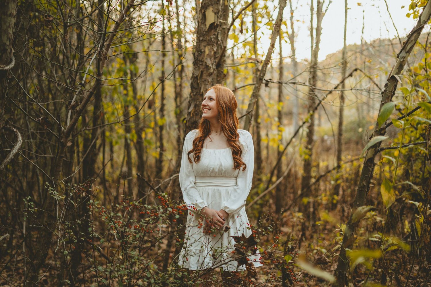 Young woman with red hair in white dress smiling among autumn trees in a forest.