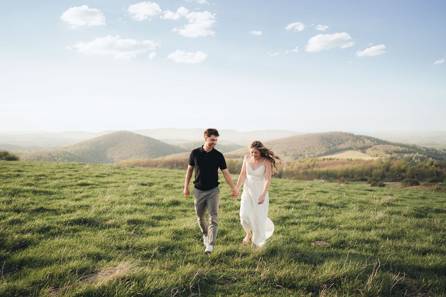 Couple walking hand-in-hand through green hilltop meadow with rolling mountain landscape and blue sky backdrop.