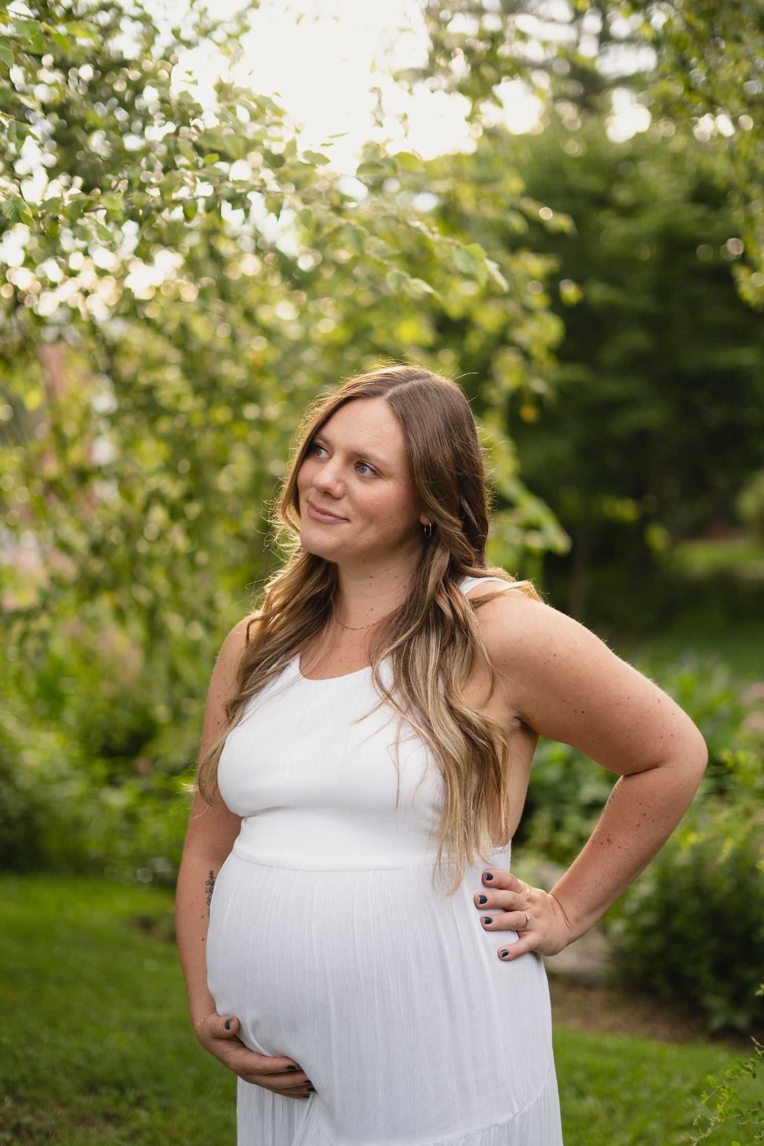 Pregnant woman in white dress smiling outdoors in a garden, hand on hip cradling her baby bump.