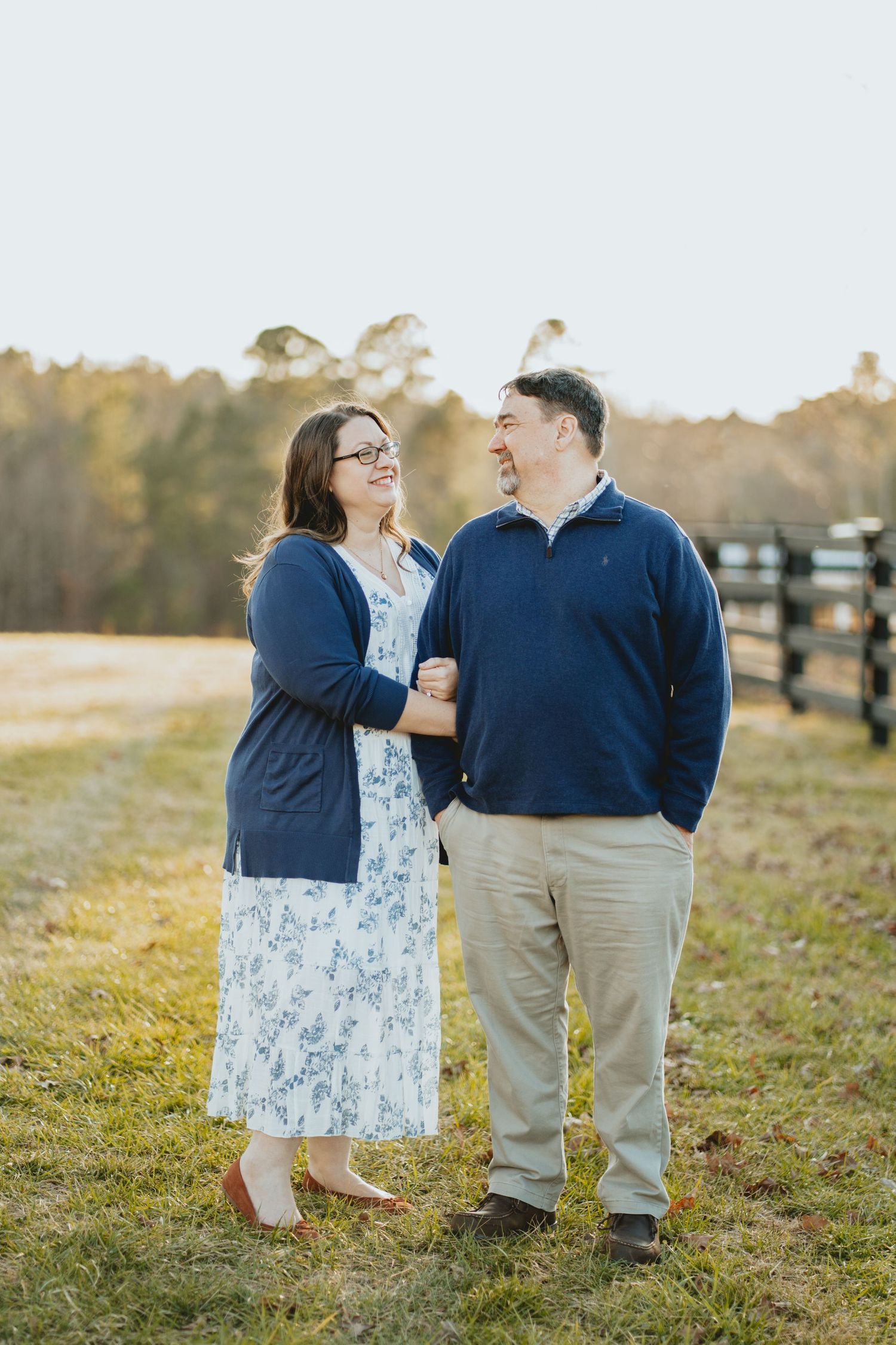 Couple laughing and holding hands in a sunny field with a wooden fence in the background.