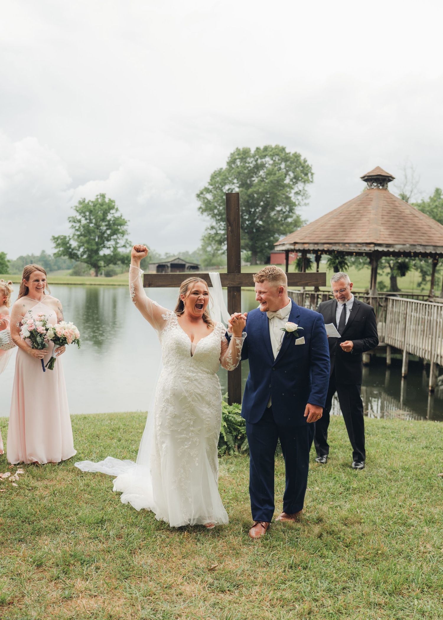 Joyful bride and groom celebrate after outdoor waterfront wedding ceremony with bridesmaids and cross backdrop.